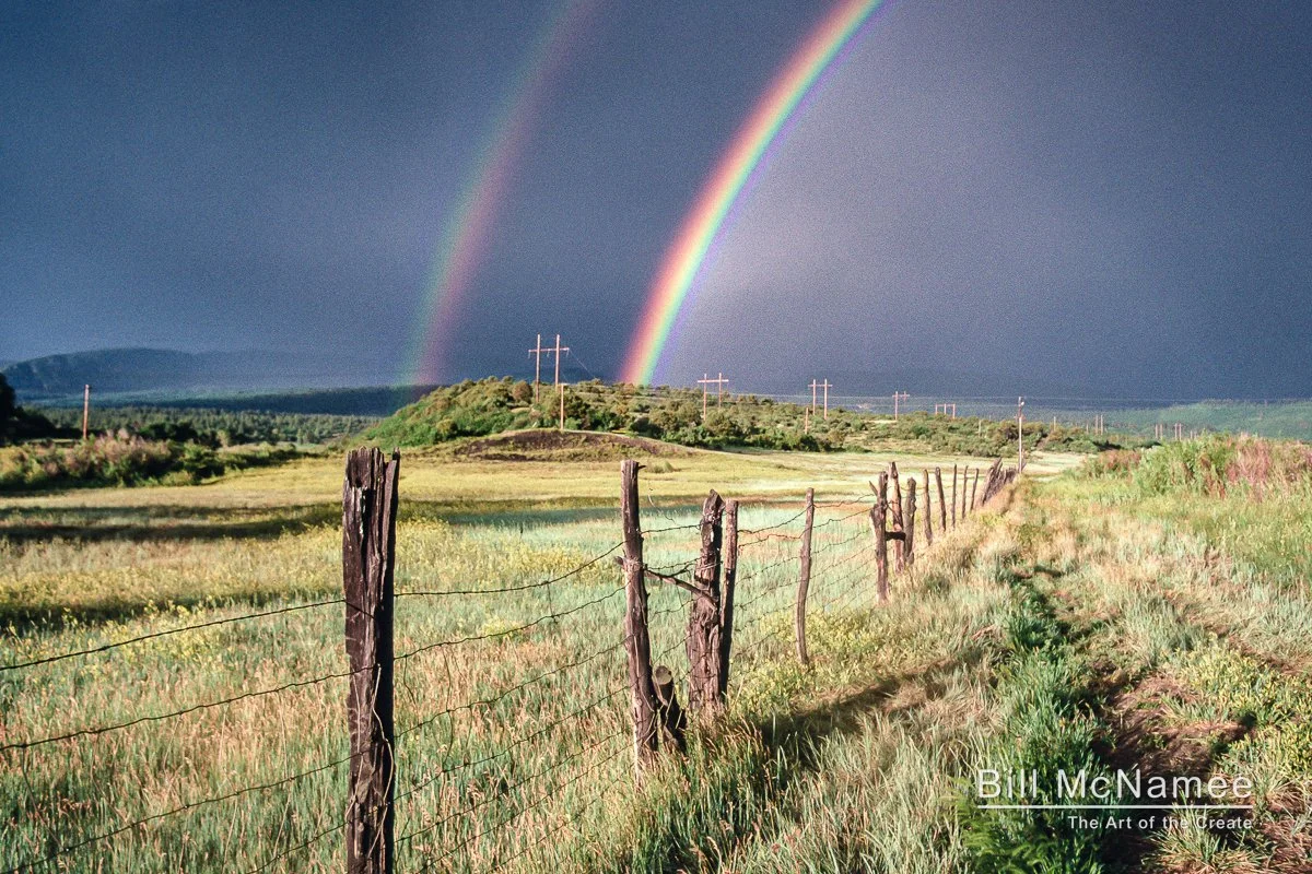 Rural landscape near Mancos, Colorado, with a weathered fence line leading toward the horizon beneath a double rainbow after a passing storm.