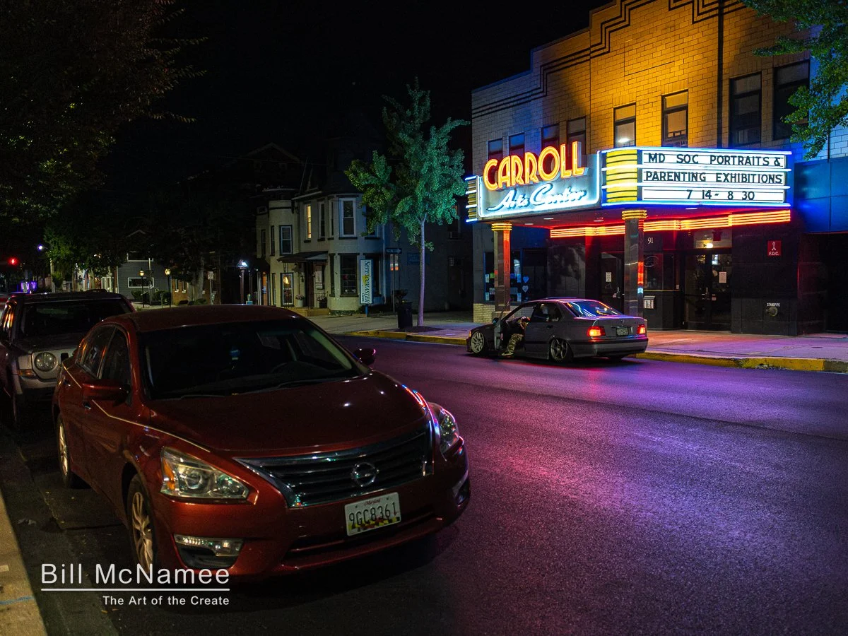 Carroll Arts Center at night with glowing marquee, parked cars, and a driver-side door open at the curb
