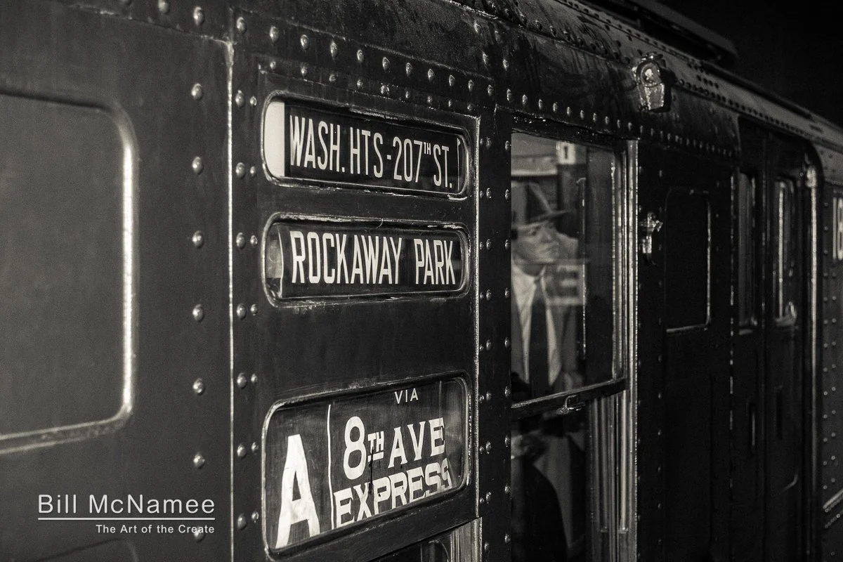 Man in suit and fedora riding the A line subway, seen through a riveted steel car window.