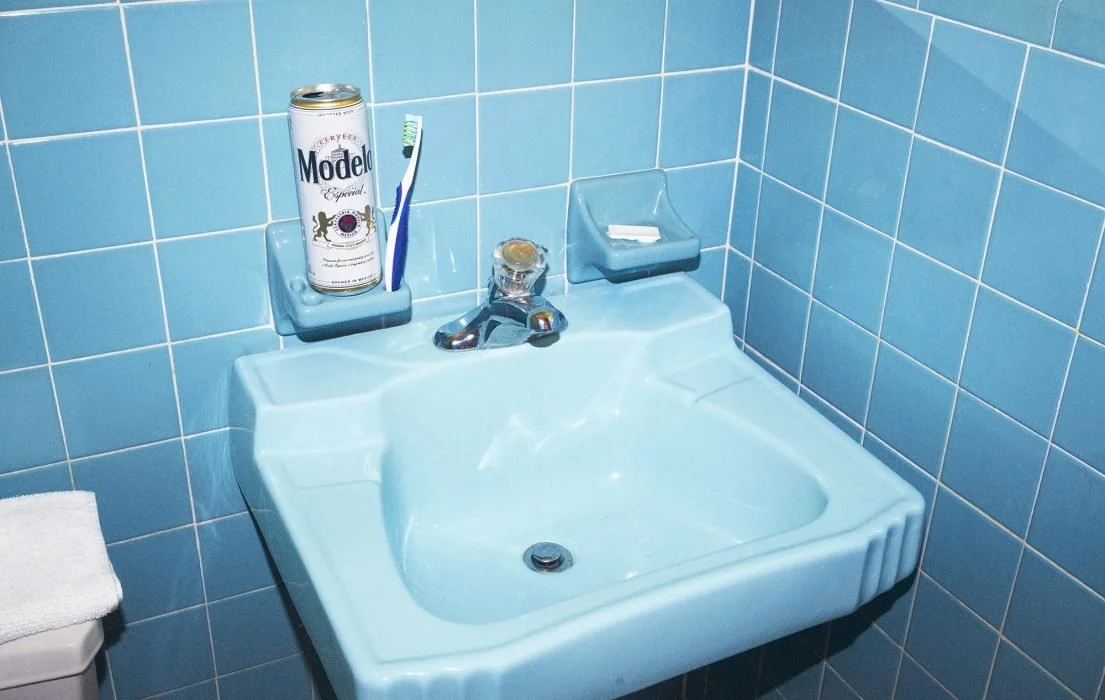 A blue bathroom sink with a soap dish, a faucet, and a can of Modelo beer, with a toothbrush and other items, mounted on a wall with blue tiles.