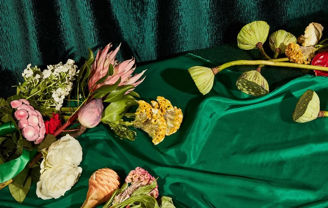An arrangement of various flowers on a green satin cloth, including pink ginger, white roses, yellow flowers, and lotus pods against a dark textured background.