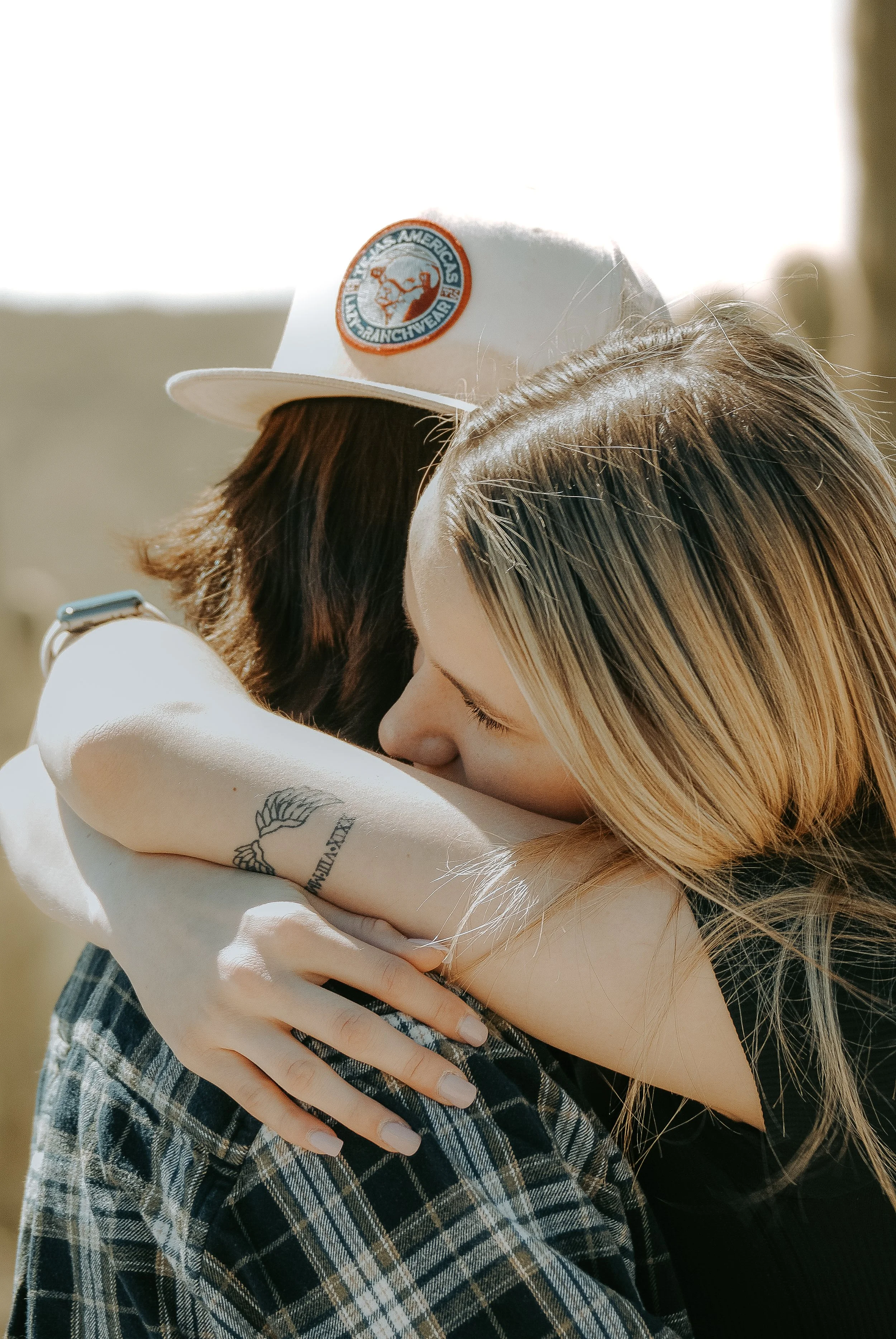 Two women hug affectionately outdoors. One has blonde hair, freckles, and a tattoo on her arm. The other has dark hair and wears a white cap with a logo.