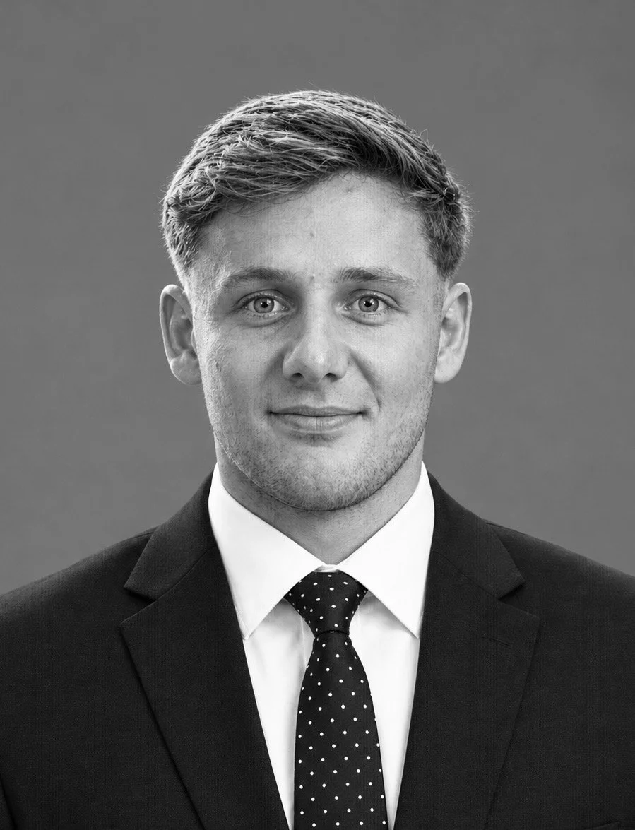 Black and white portrait of a young man with short, styled hair, wearing a collared shirt and a polka dot tie, smiling slightly against a dark background.