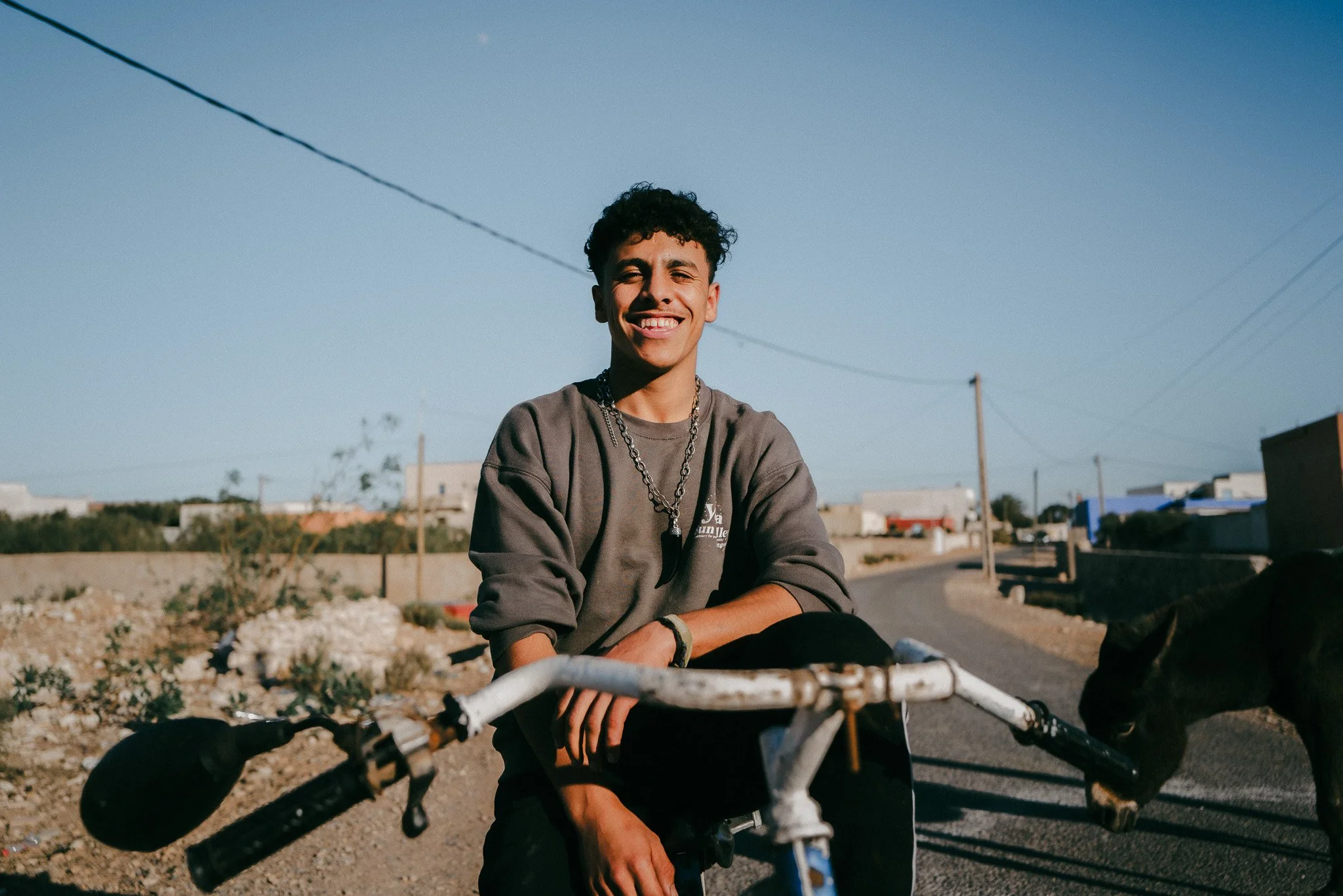 Ein junger Mann mit dunklen, lockigen Haaren sitzt auf seinem Fahrrad, lächelt und trägt eine Kette. Im Hintergrund ist eine Straße in einem ländlichen Gebiet mit wenigen Gebäuden und einem Hund zu sehen.