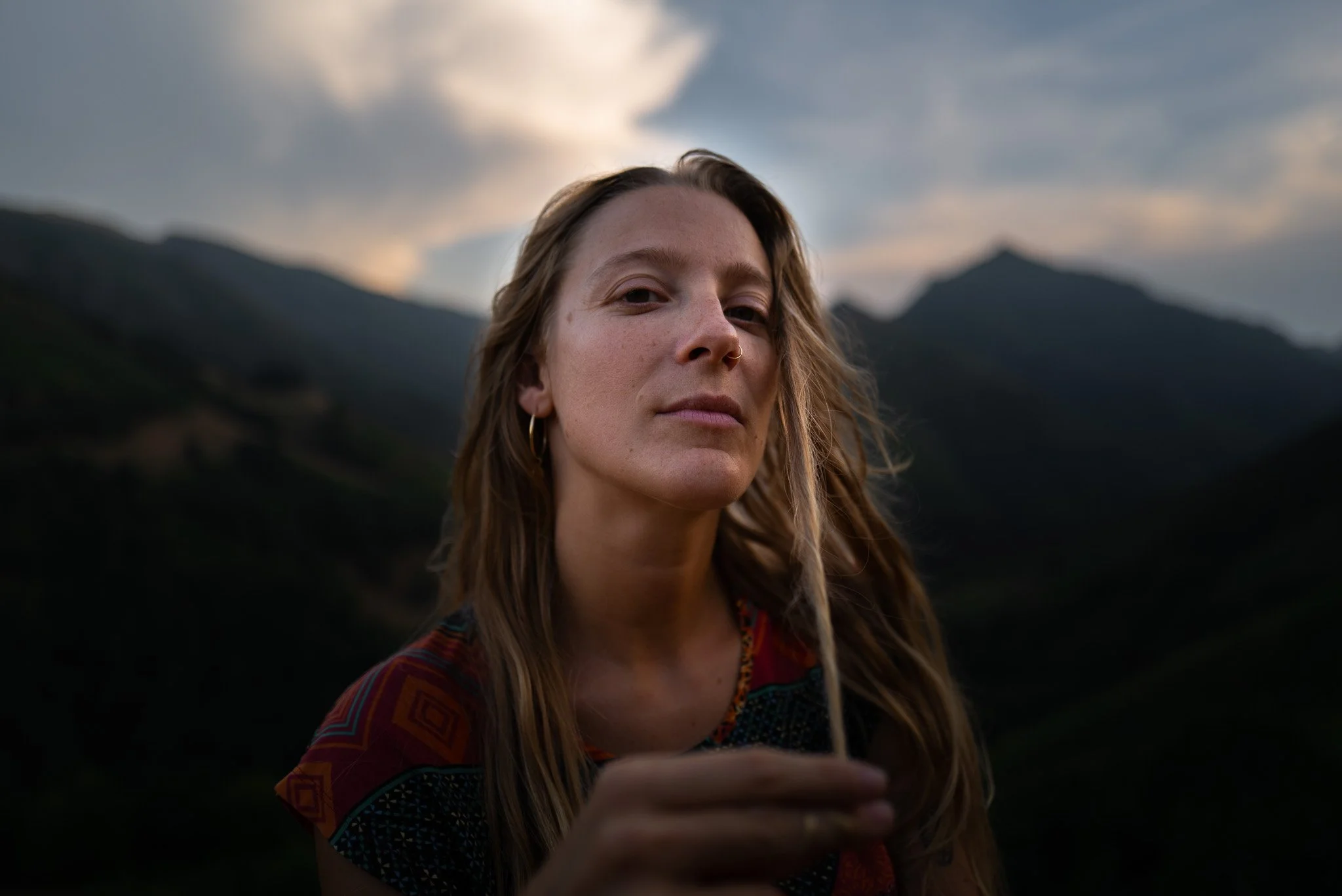 Nahaufnahme einer jungen Frau mit langen, gewellten Haaren, die in einer Berglandschaft bei Sonnenuntergang oder Dämmerung steht, hält ein Gras oder eine Pflanze in der Hand.