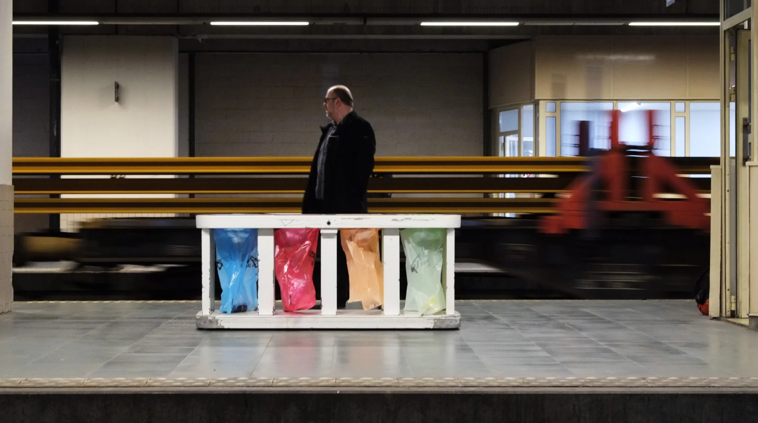 Ein Mann steht auf einem Bahnsteig und blickt nach unten. Im Hintergrund ist eine rote Straßenbahn, die in Bewegung ist. Vor dem Mann befindet sich eine weiße Absperrung mit vier Beuteln in verschiedenen Farben (blau, rot, beige, grün). Das Bahnsteig