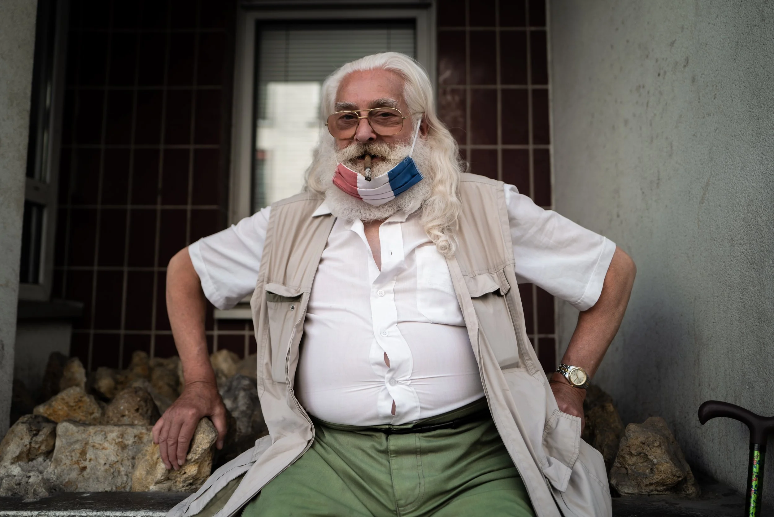 Älterer Mann mit weißen locked Haaren, Brille, Tatoo, und Schnurrbart, sitzt auf einer Bank vor einer Hauswand, trägt eine Maske mit französischer Flagge, trägt ein weißes Hemd, eine Weste und eine Uhr.