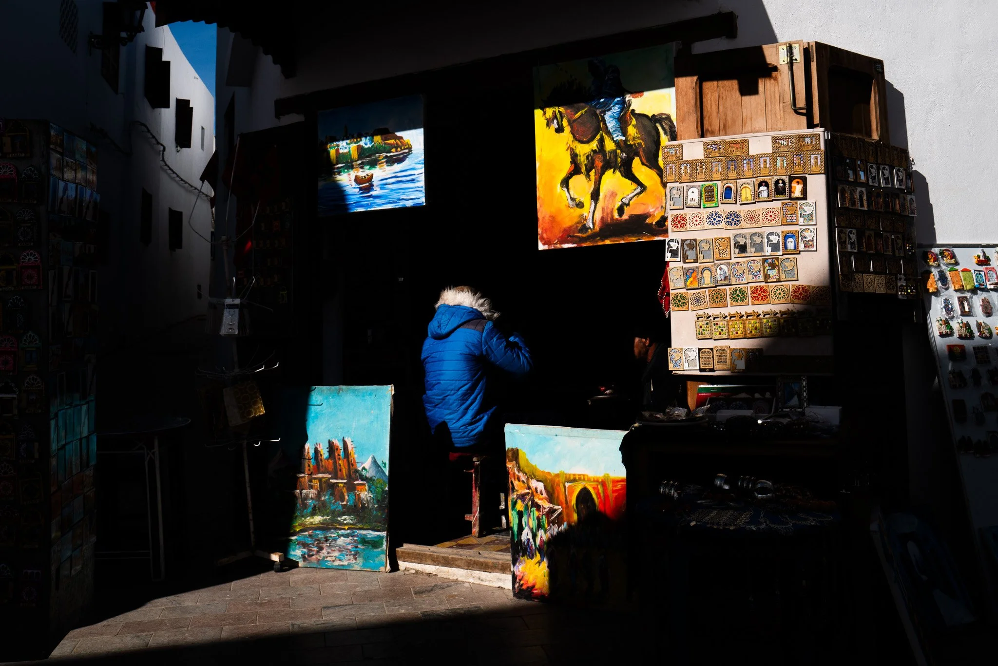 Ein Straßenkünstler verkauft bunte Gemälde und Kunsthandwerk in einer dunklen Markthalle, beleuchtet von Sonnenlicht durch eine Öffnung.