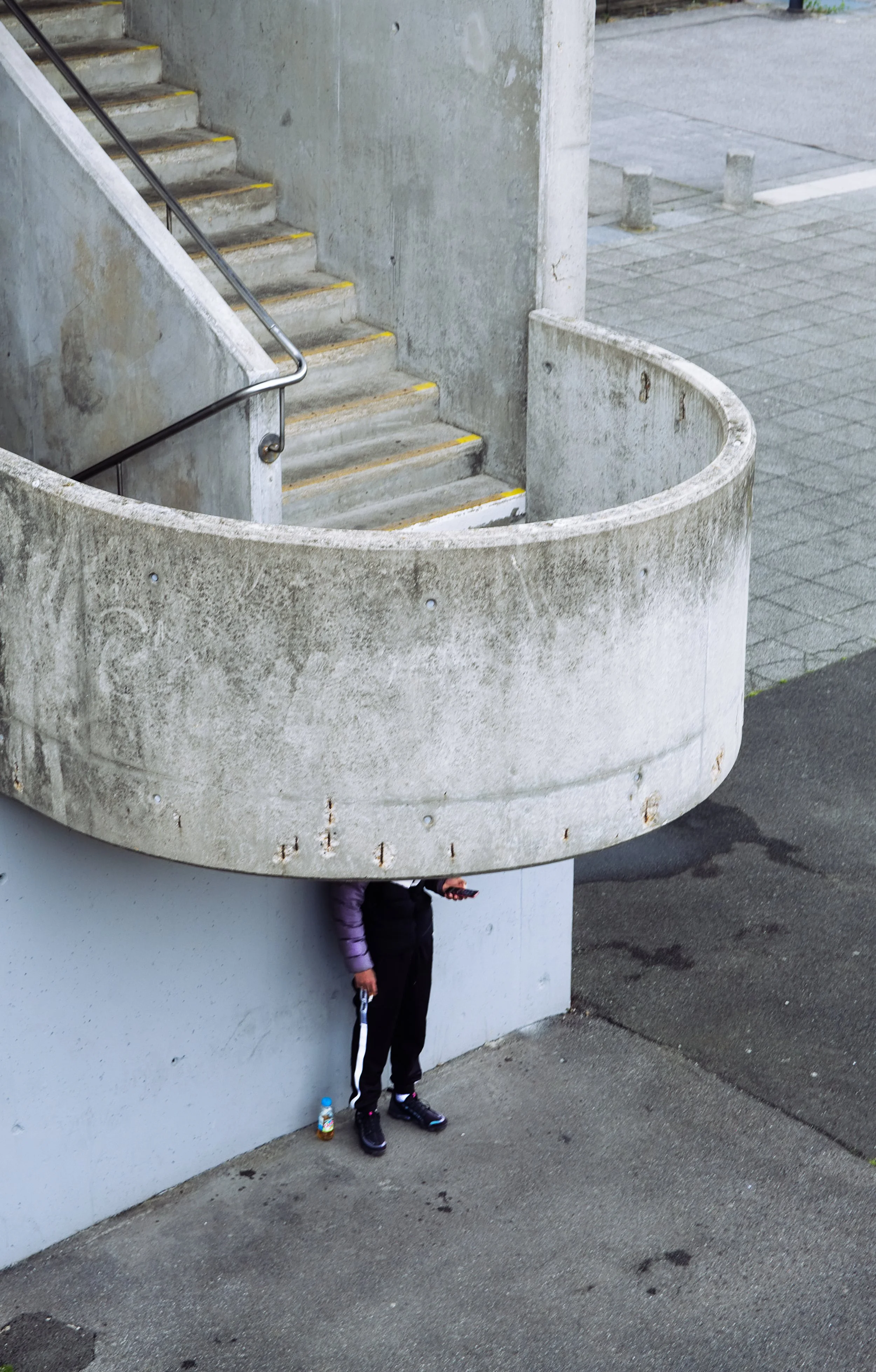 Ein Mensch steht mit dem Kopf unter einer großen, runden Betontreppe, die an einer Wand angebracht ist, und hält ein Smartphone in der Hand, während er auf dem Boden steht. Neben dem Menschen steht eine Wasserflasche.