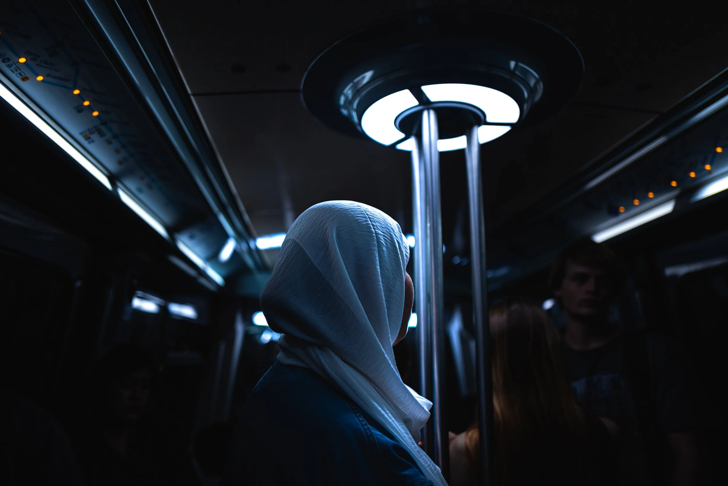 Menschen in einem dunklen Bus mit blauer Beleuchtung, eine Person trägt ein weißes Kopftuch, andere stehen or sitzen im Hintergrund.
