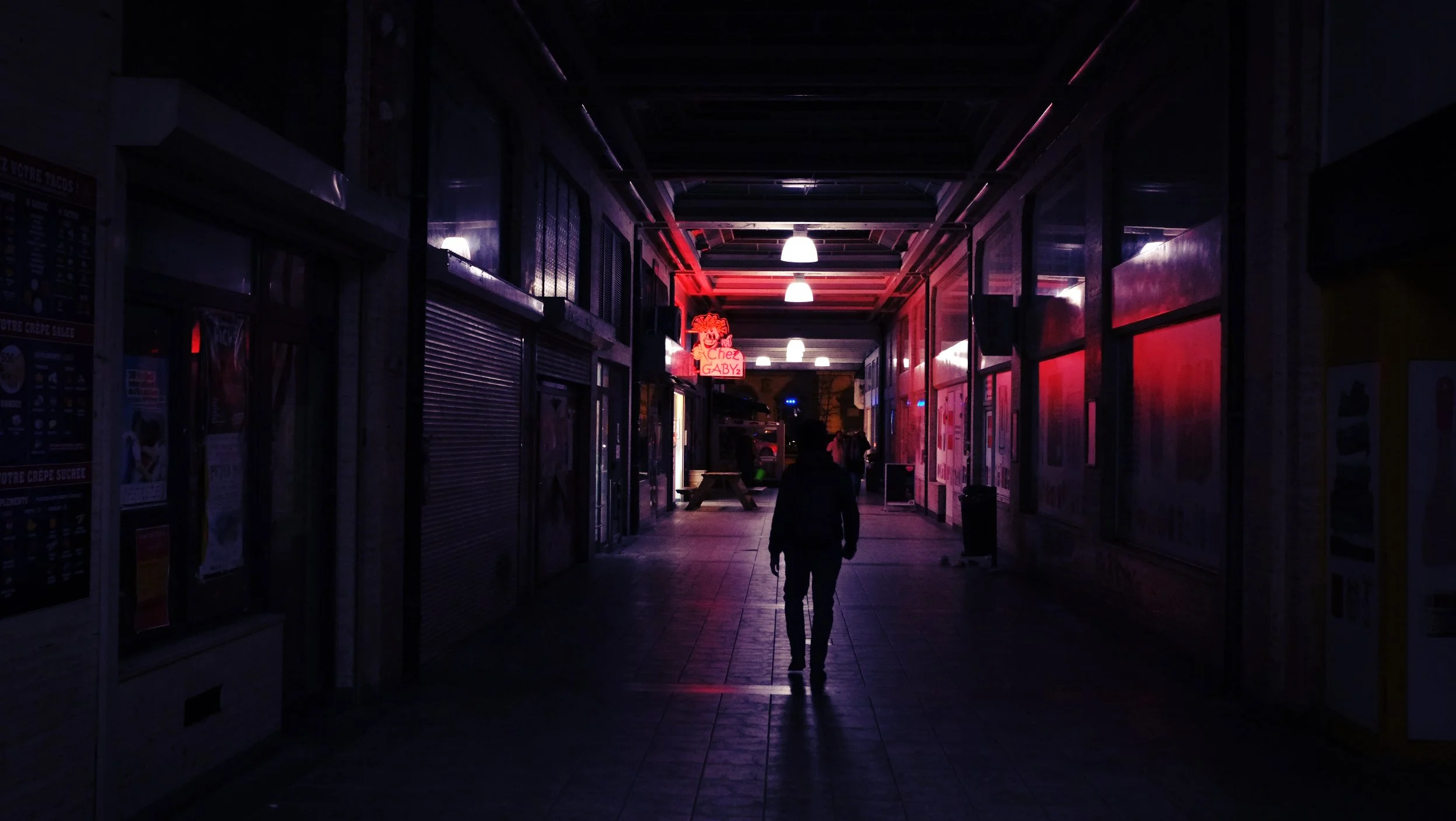 Ein dunkler Korridor mit Neonlichtern und Schaufenstern, eine einzelne Person geht in Richtung Ende.