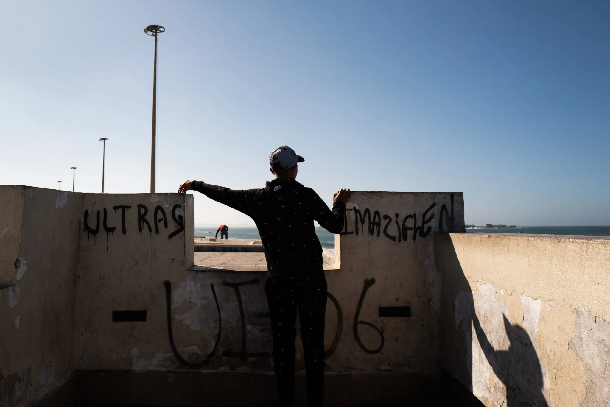 Junge steht an einer Sonnenbrille, trägt schwarze Kleidung und schaut auf den Strand. Graffiti auf der Wand, Blick aufs Meer bei Sonnenuntergang, ruhige Atmosphäre.