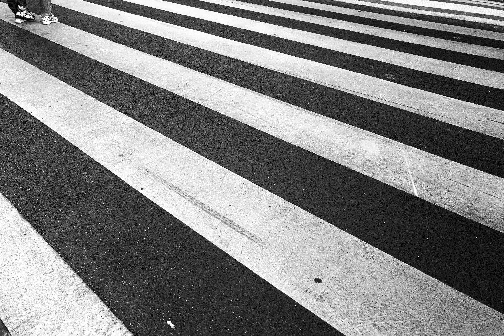 Schwarz-weiße Zebrastreifen auf der Straße, mit zwei Personen, die an der oberen linken Ecke vorbeigehen.