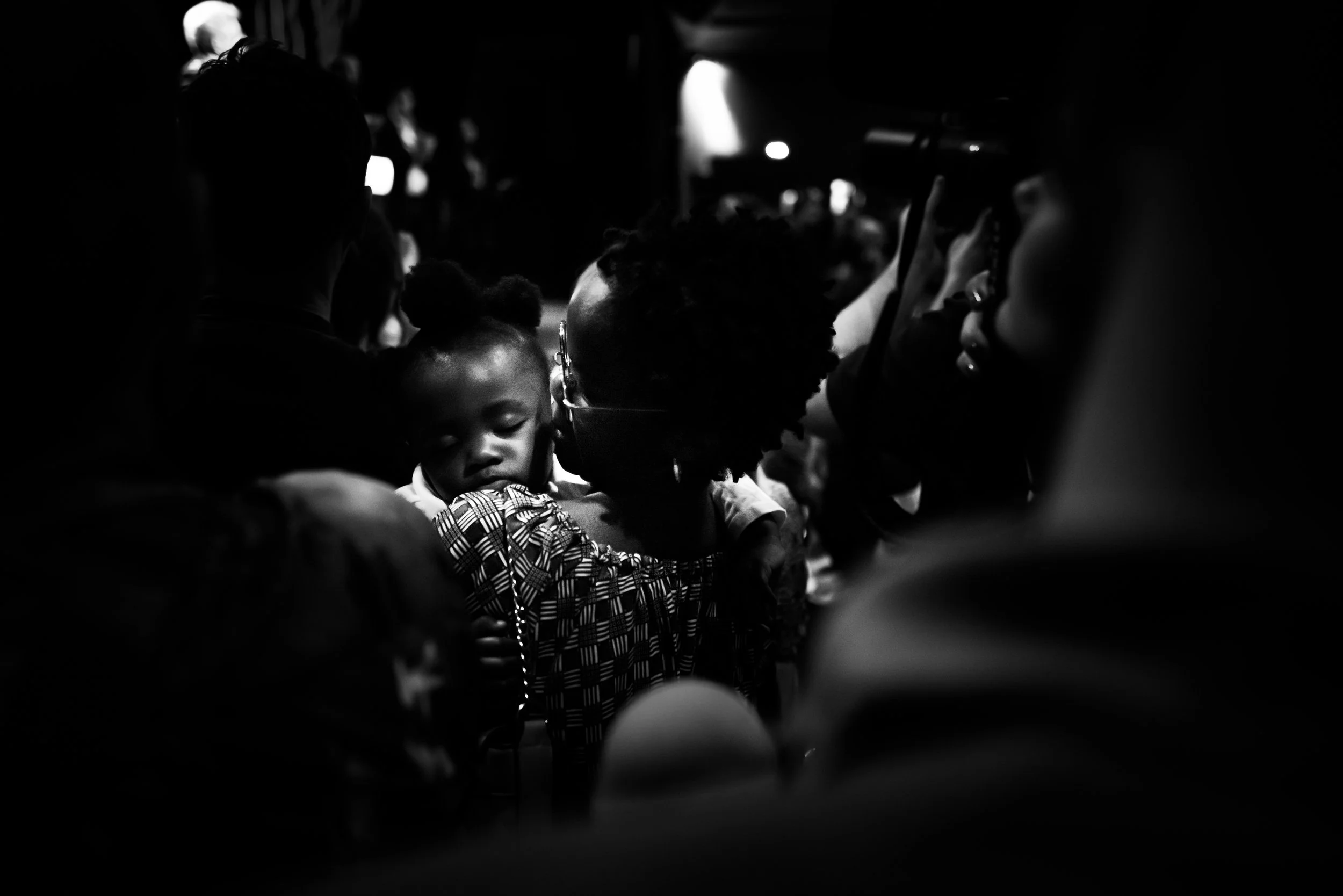 Schwarzes und weißes Foto von einer Menschenmenge, im Mittelpunkt ein schlafendes Kind auf einer Schulter einer erwachsenen Person, umgeben von weiteren Menschen in dunklem Hintergrund.