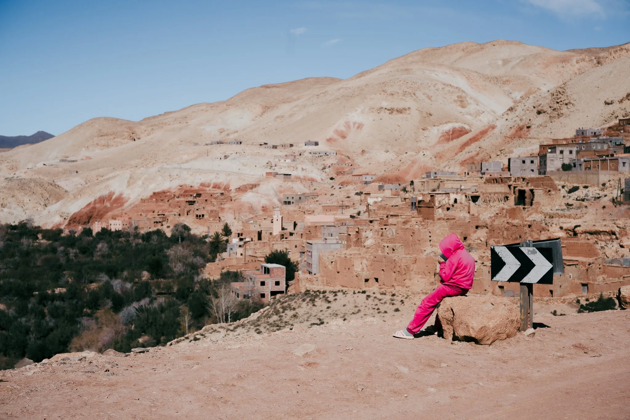 Ein Kind in pinker Kleidung sitzt auf einem Stein am Straßenrand in einer trockenen Berglandschaft mit einer Stadt im Hintergrund, umliegende Hügel und Gebäude sichtbar.