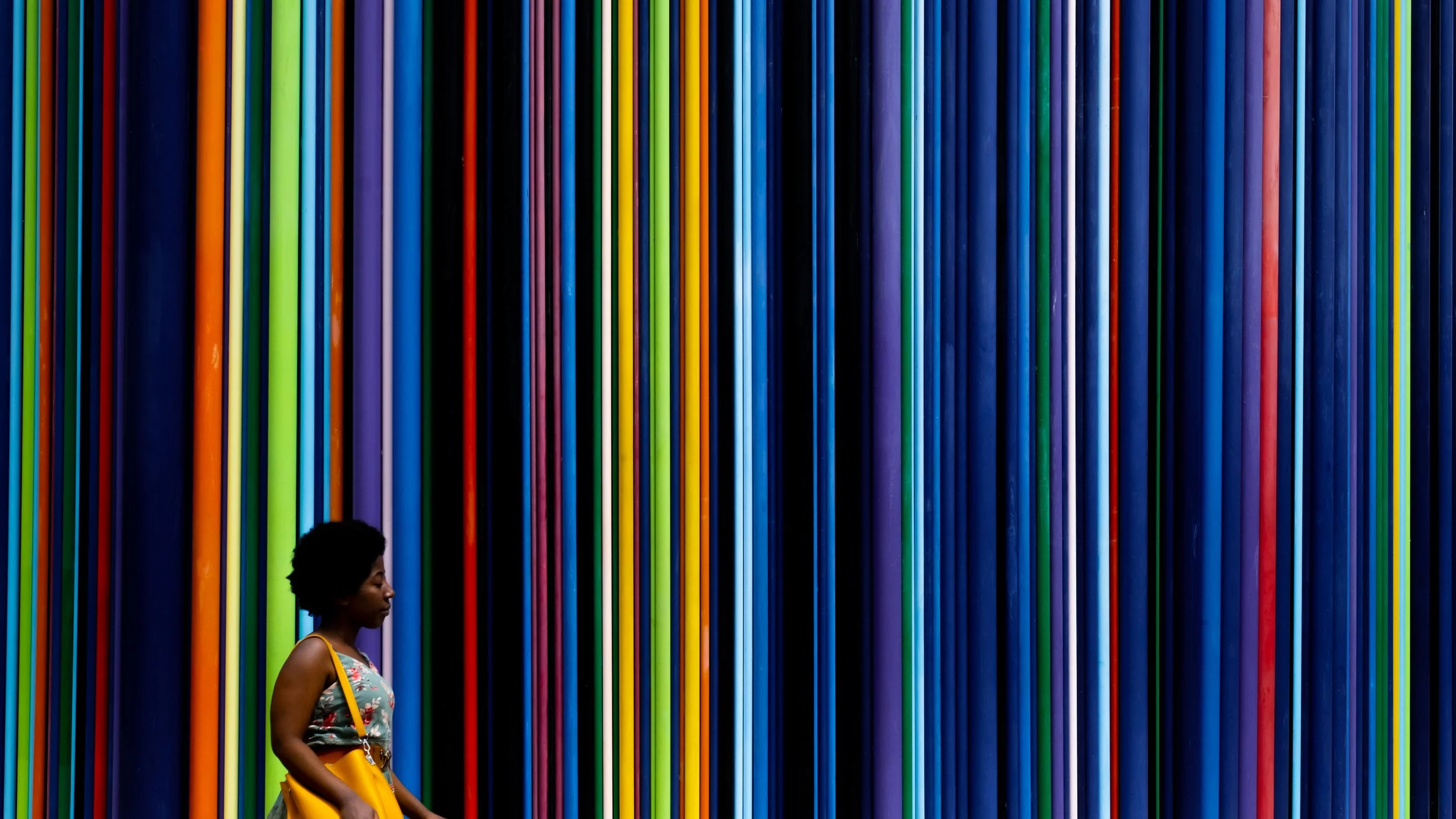 Eine Frau mit Afro-Haar, die eine gelbe Tasche trägt, steht vor einer Wand mit vertikalen, bunten Streifen in Dunkelblau, Schwarz, Orange, Grün, Hellblau, Lila und Blau.