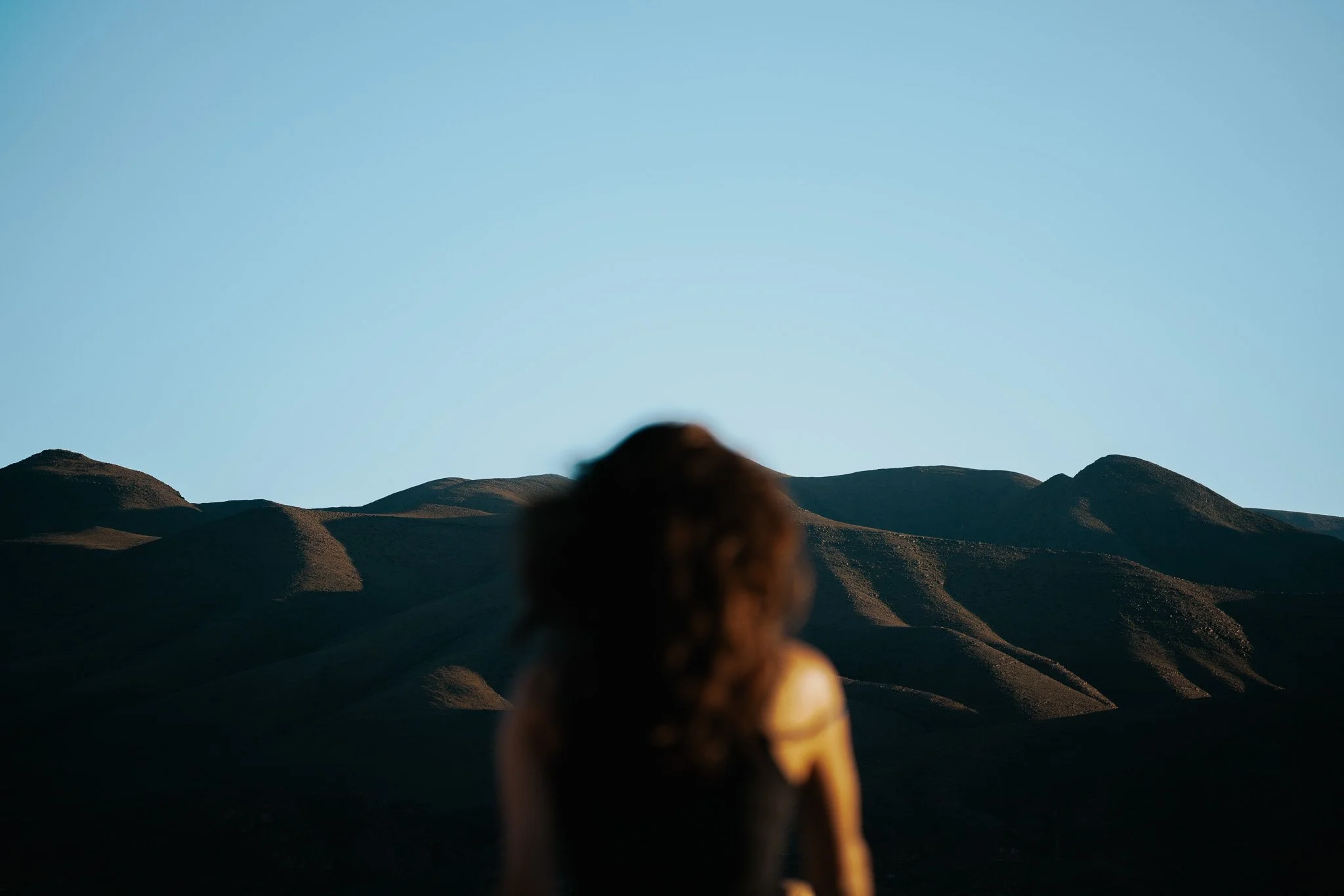 Eine Person mit dunklen Haaren, die in die Ferne blickt, vor einer Landschaft mit Hügeln und klarem blauen Himmel.