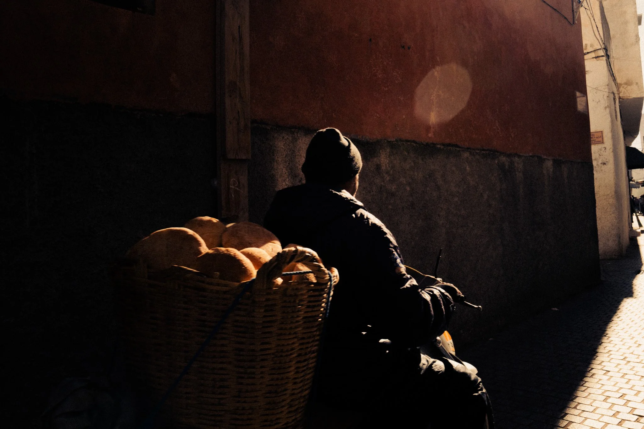 Eine Person sitzt an einer Hauswand, im Hintergrund scheint die Sonne auf den Bürgersteig, mit einem Korb voller Brötchen auf dem Rücken.