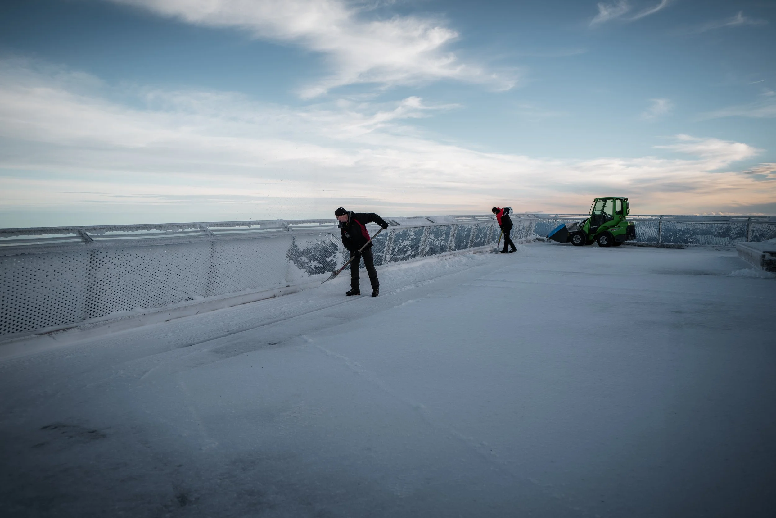 Mitarbeiter reinigen die Dachfläche eines Gebäudes im Winter mit Schneeschaufeln und einem Schneefräser, während die Wolken am Himmel zu sehen sind.