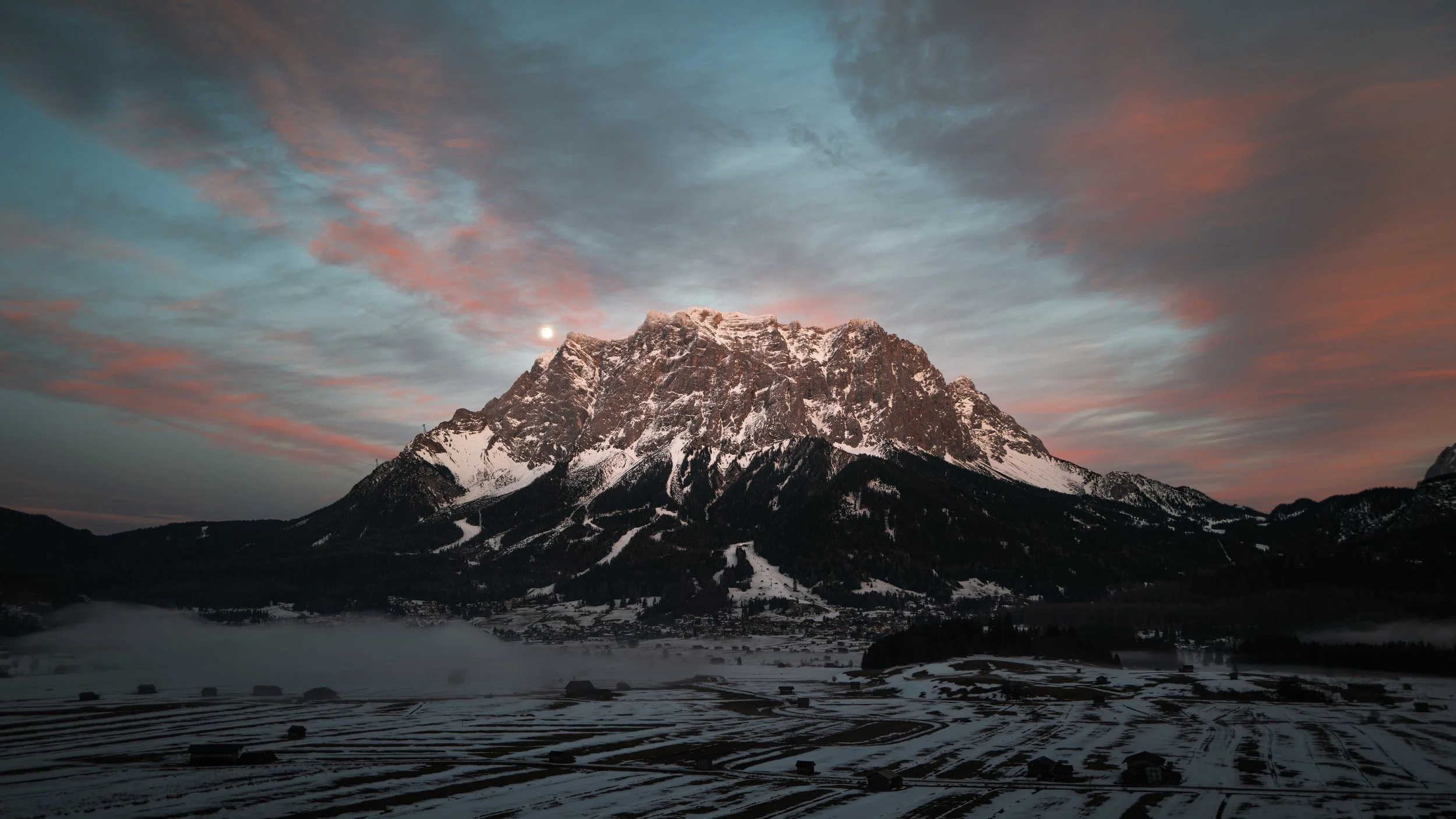 Berg im Schnee bei Sonnenuntergang mit lila und rosa Wolken am Himmel, verschneite Felder im Vordergrund.