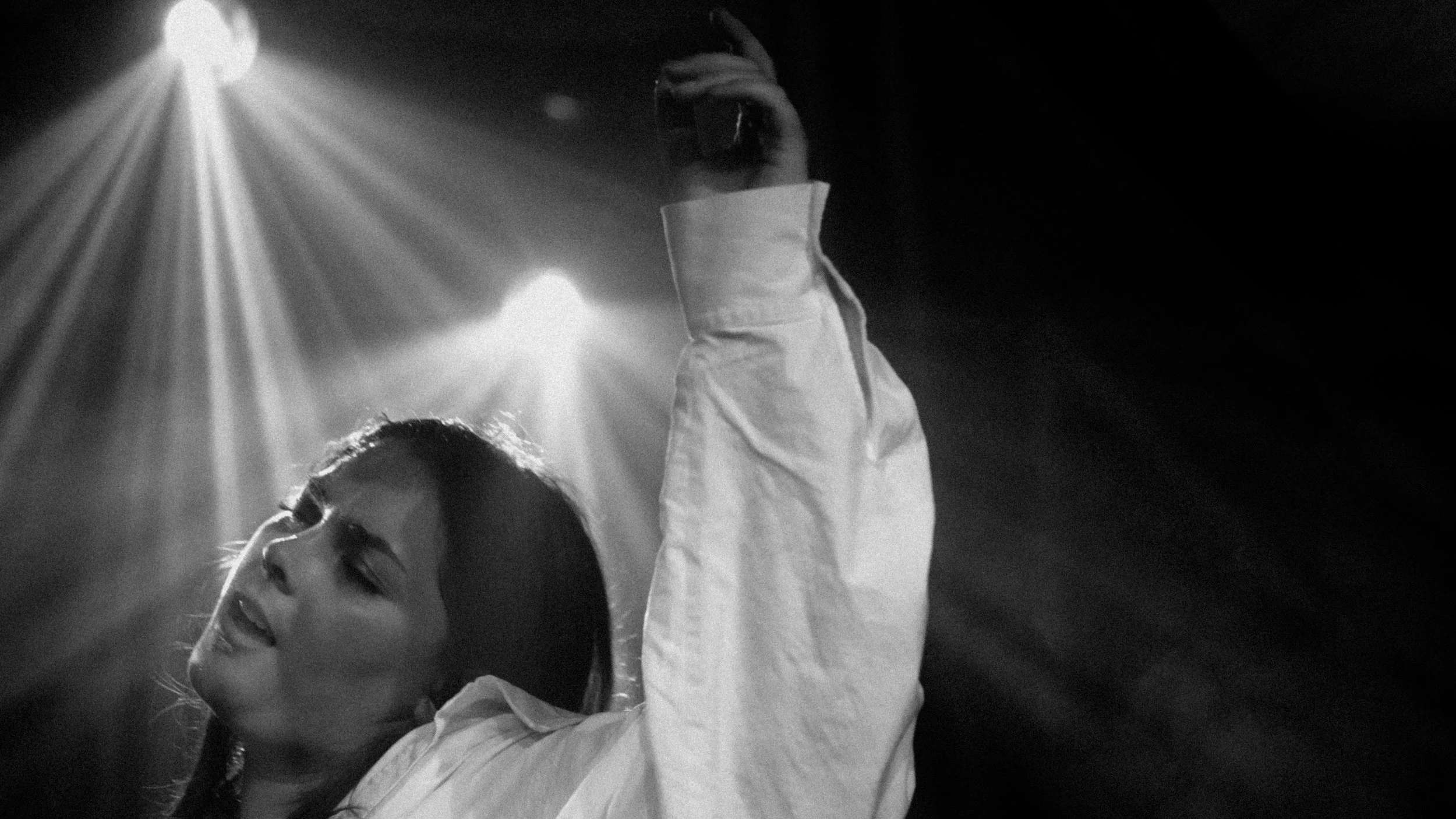 Schwarz-weiß-Foto eines Mädchens mit geschlossenen Augen, das mit ausgestrecktem Arm nach oben greift, vor dunklem Hintergrund mit Scheinwerferlicht.