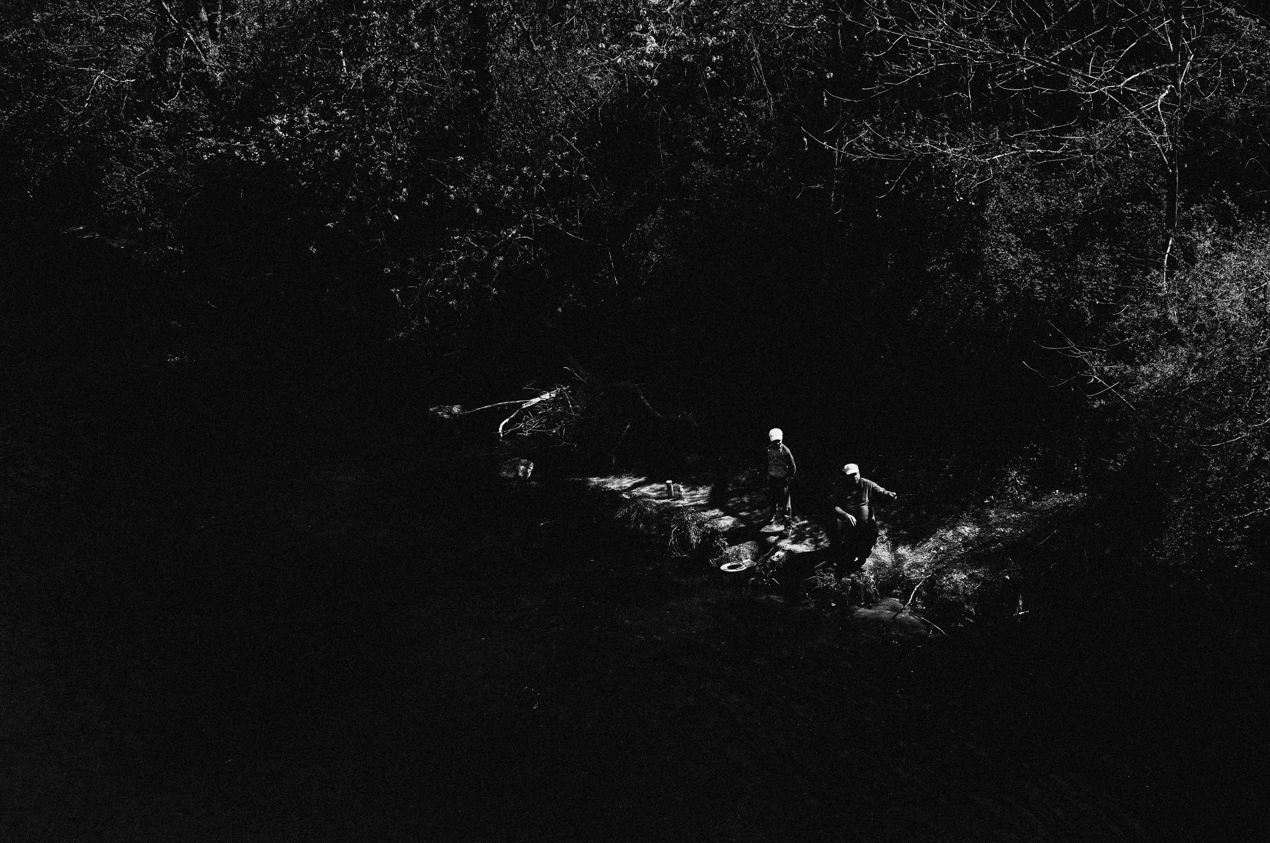 Zwei Menschen beim Wasserholen aus einem Fluss in einem dunklen Wald, schwarz-weiß.