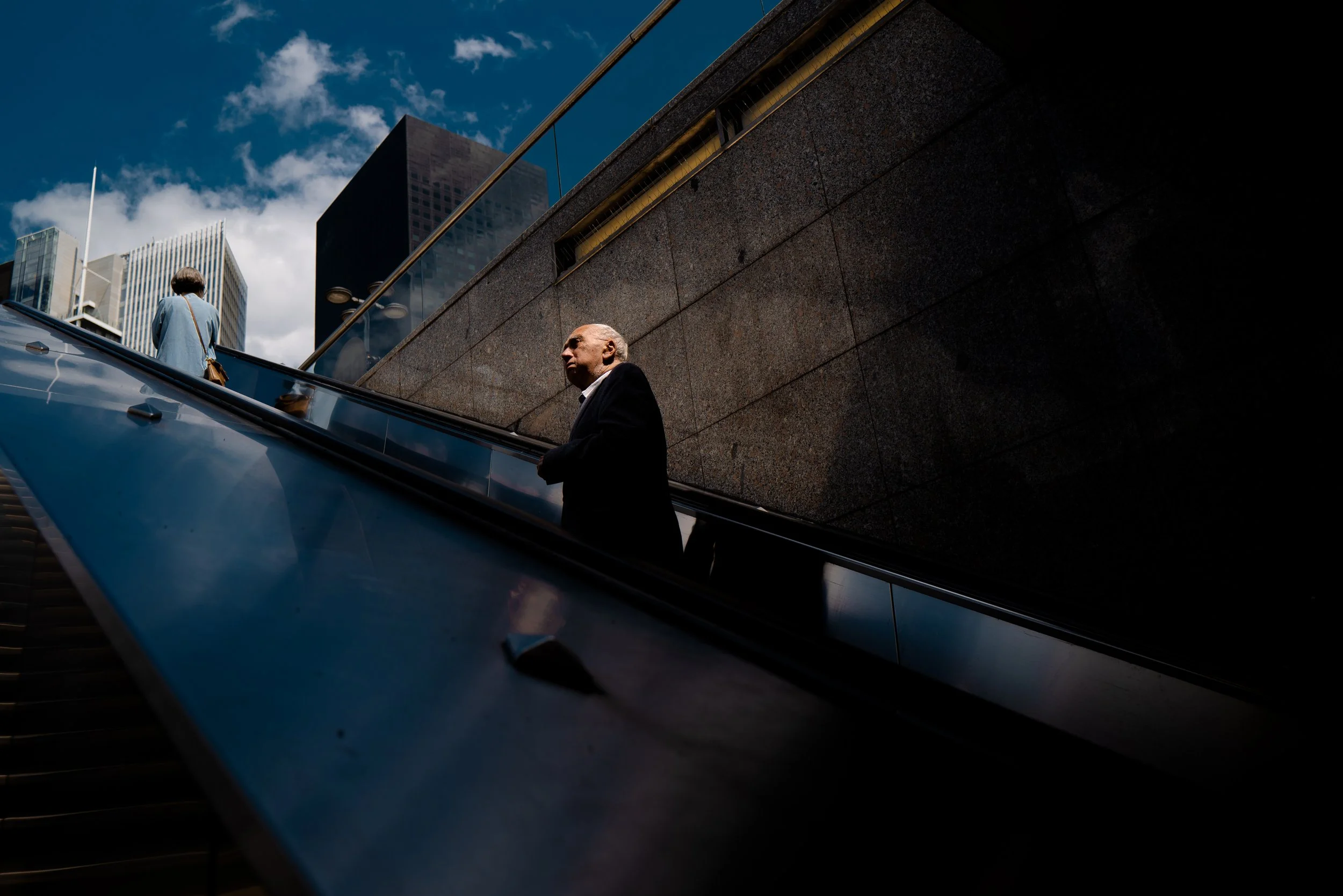 Ein Mann in dunklem Anzug steht auf einer Rolltreppe in einer modernen Stadt mit hohen Gebäuden bei bewölktem Himmel.