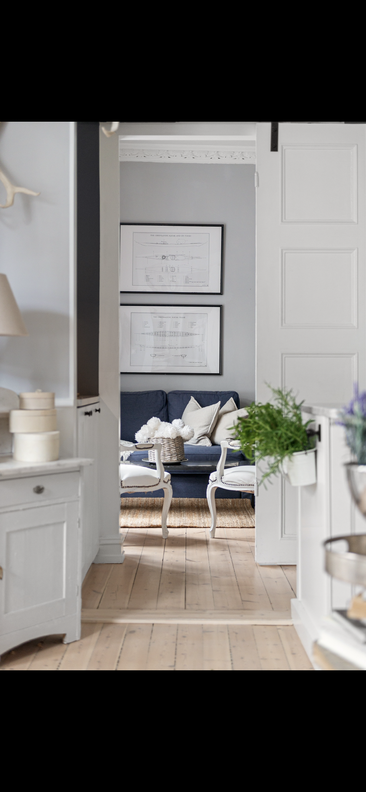 Living room with navy sofa, framed ship blueprints on light gray wall, white chairs, and decorative pillows, viewed through a doorway from an adjacent room with white cabinets and wooden floor.