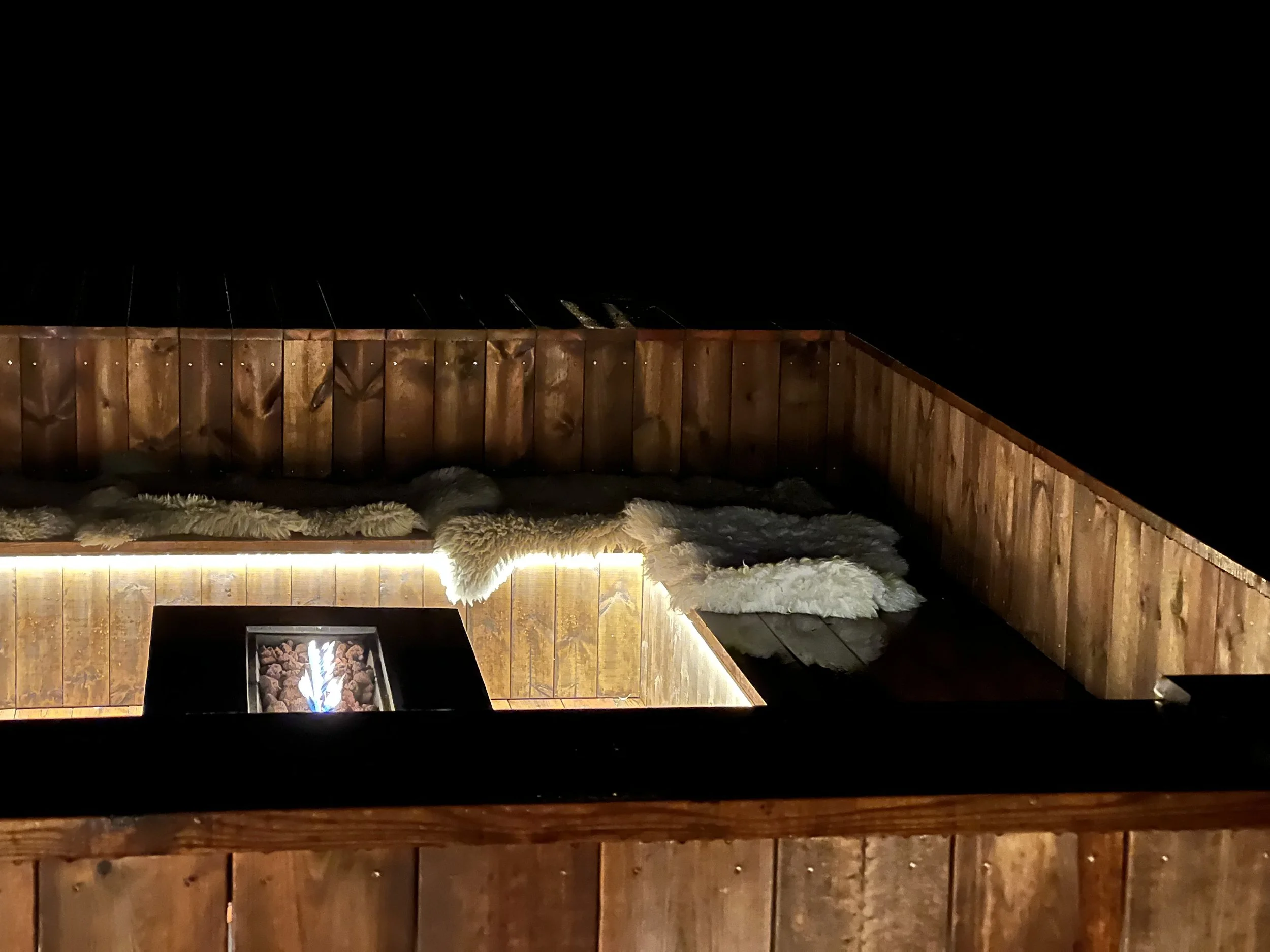 Nighttime outdoor scene featuring a wooden deck with a built-in fire feature, surrounded by a wooden privacy fence. Plush white and gray blankets are draped over the deck's edge. Darkness surrounds the area.