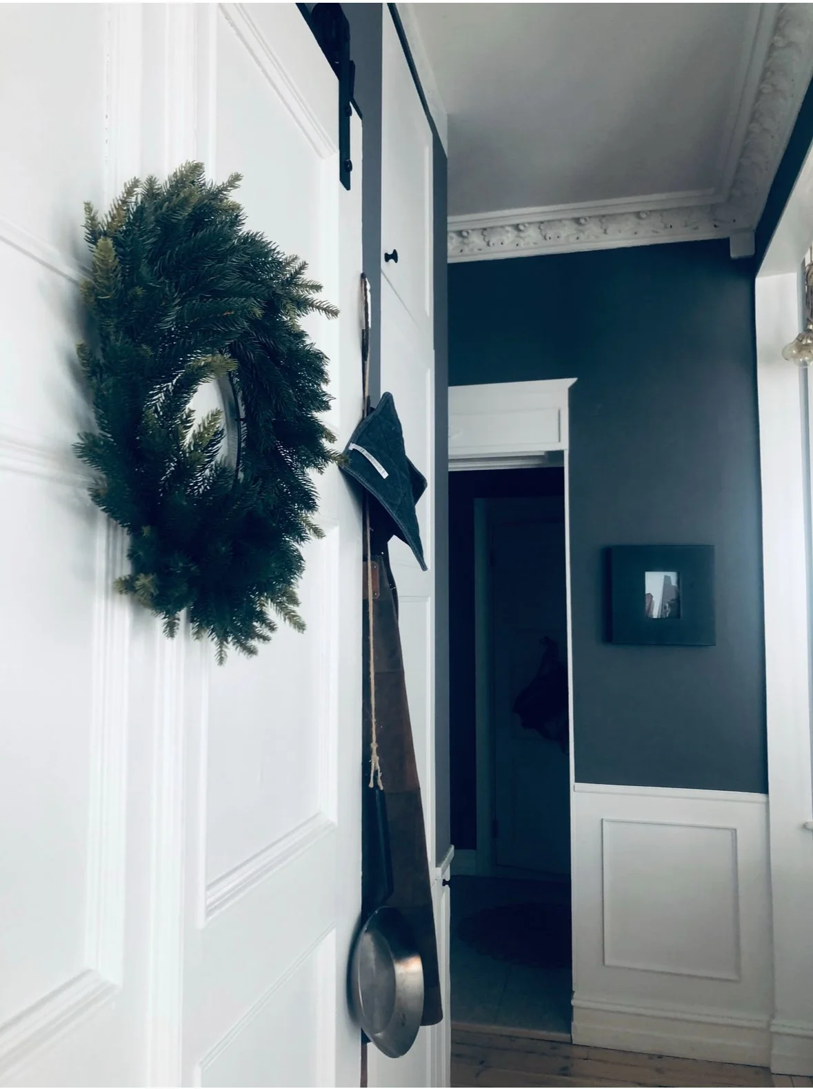 A white door with a green Christmas wreath hanging on it. A black oven mitt and a wooden handle of a long cooking spoon are hanging on hooks on the door. The background shows a hallway with dark blue walls and white trim, and a picture frame on the wall.