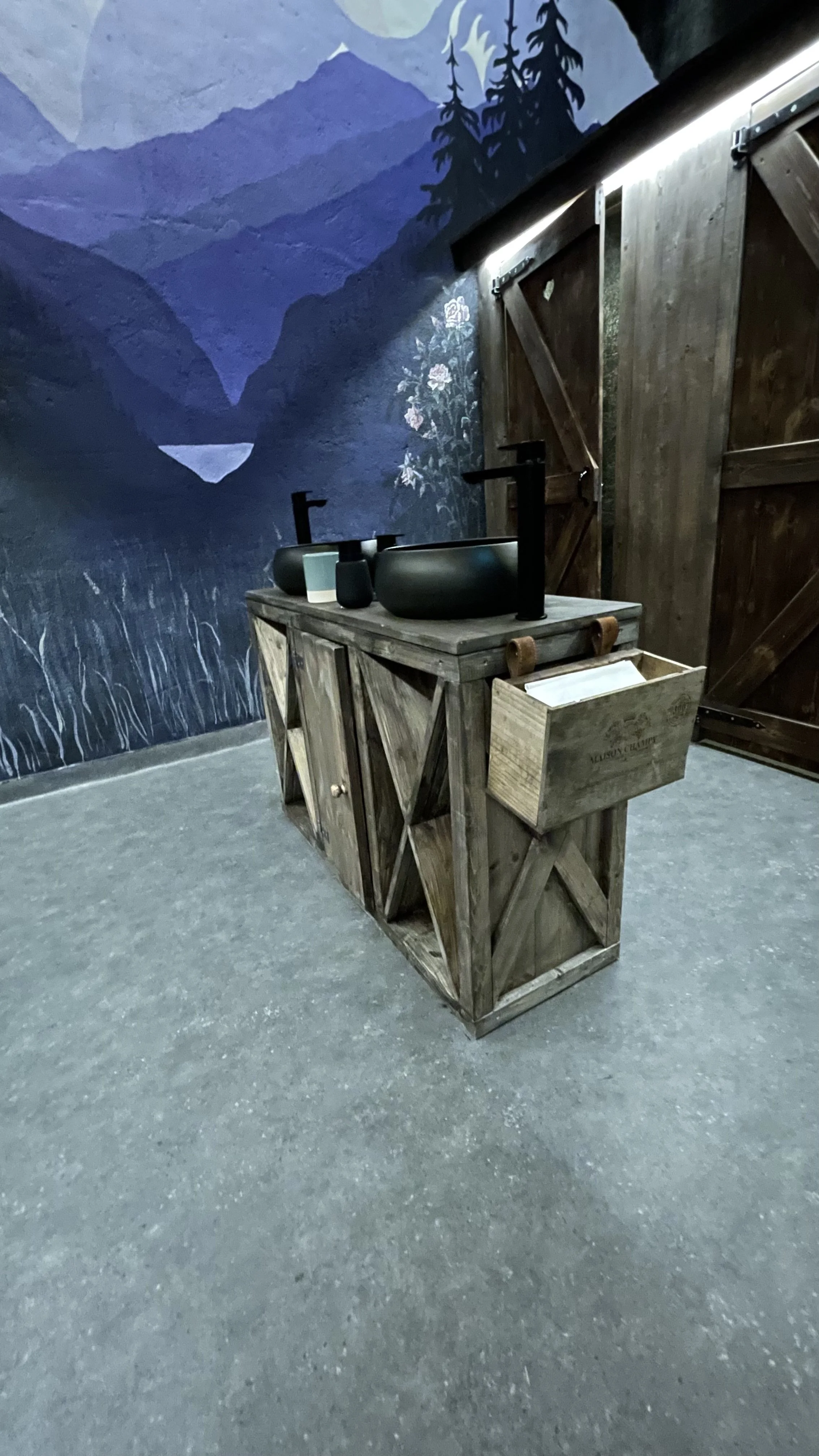 A rustic wooden vanity with two black vessel sinks and black faucets, set against a mountain landscape mural on a wall.