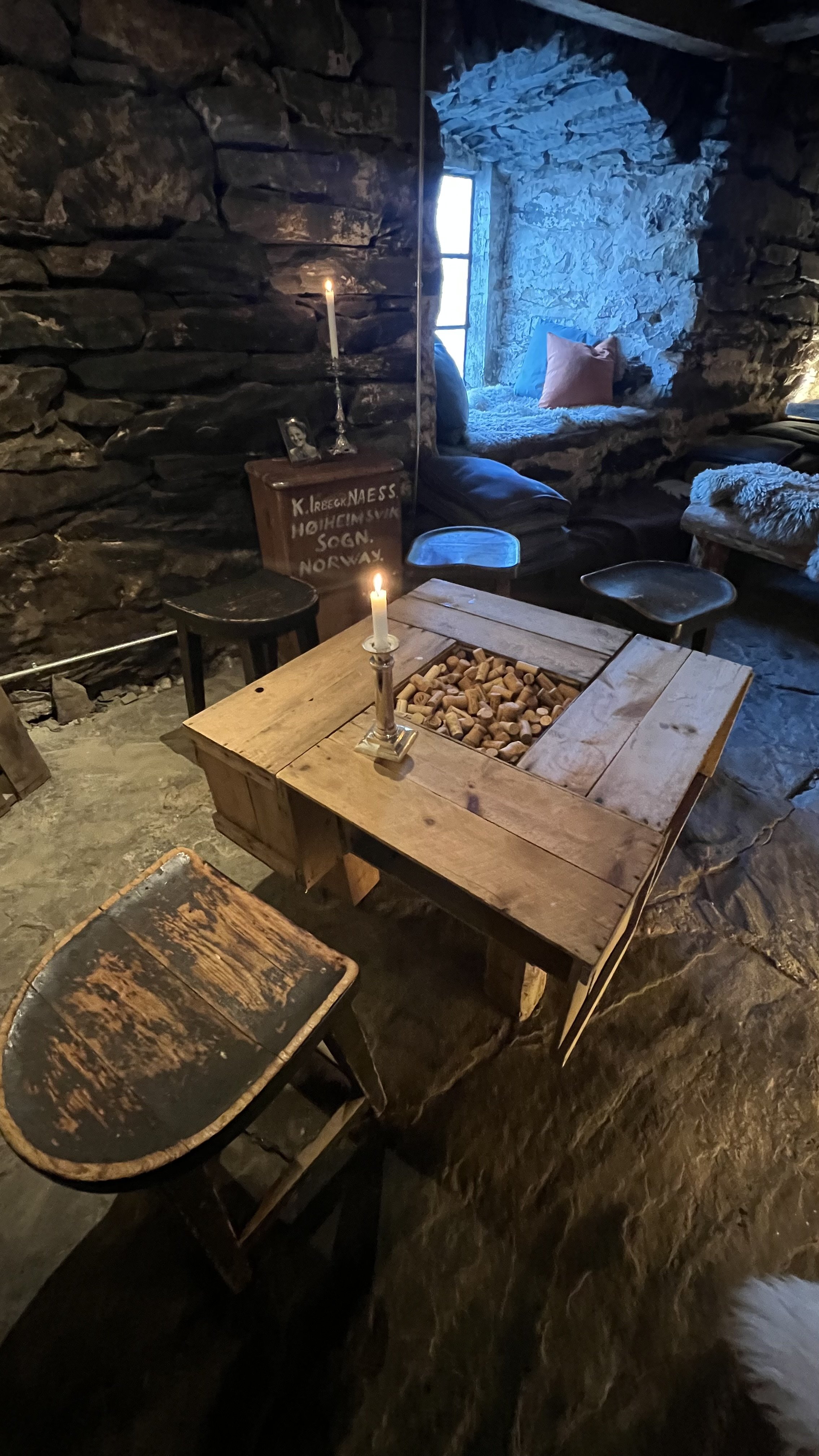 A cozy rustic room with stone walls, a wooden table with corks, candles, and a small window seat with pillows, illuminated by natural light from the window.