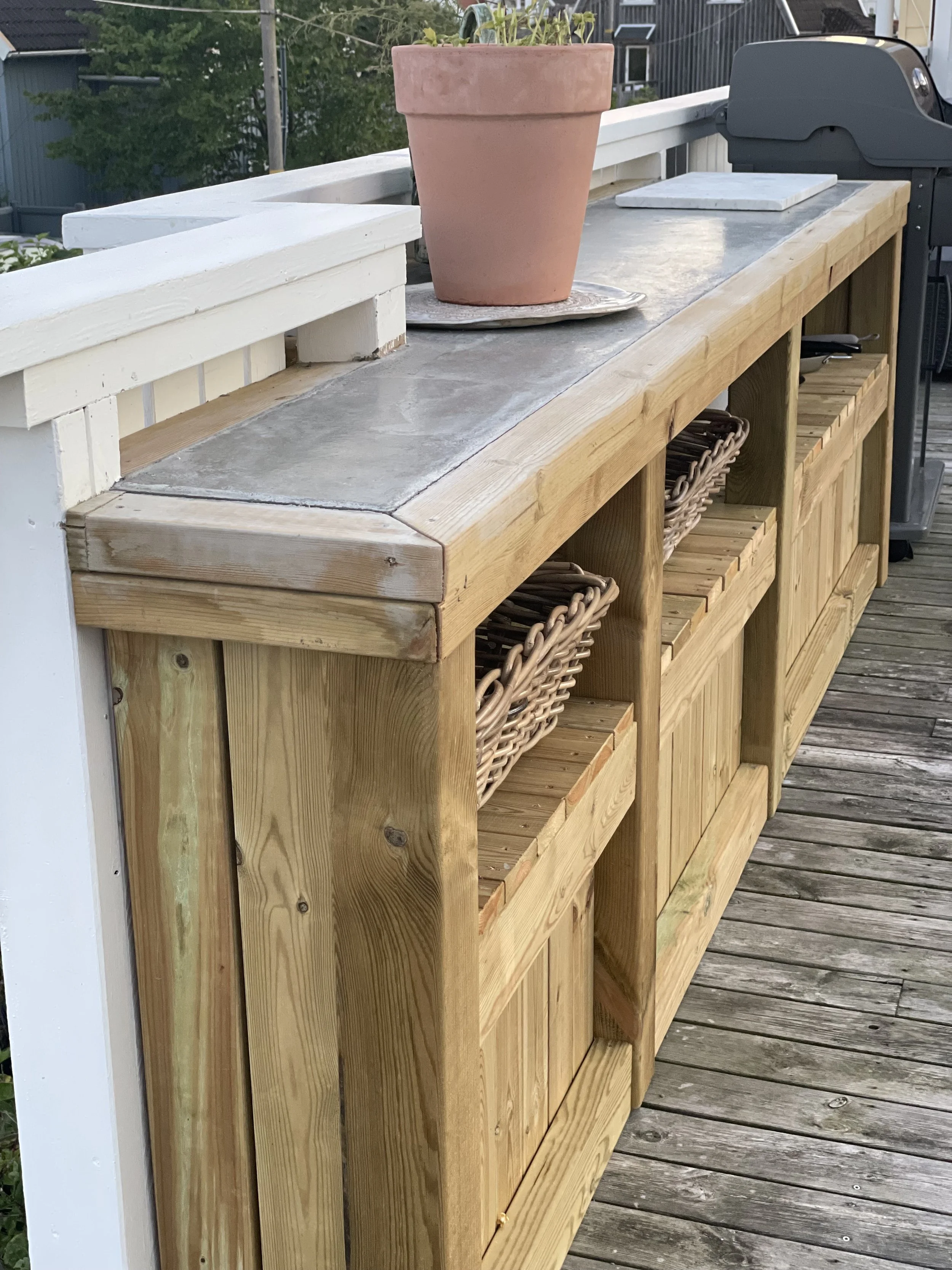 Wooden outdoor sideboard with woven baskets and a potted plant on top, on a wooden deck with a grill in the background.