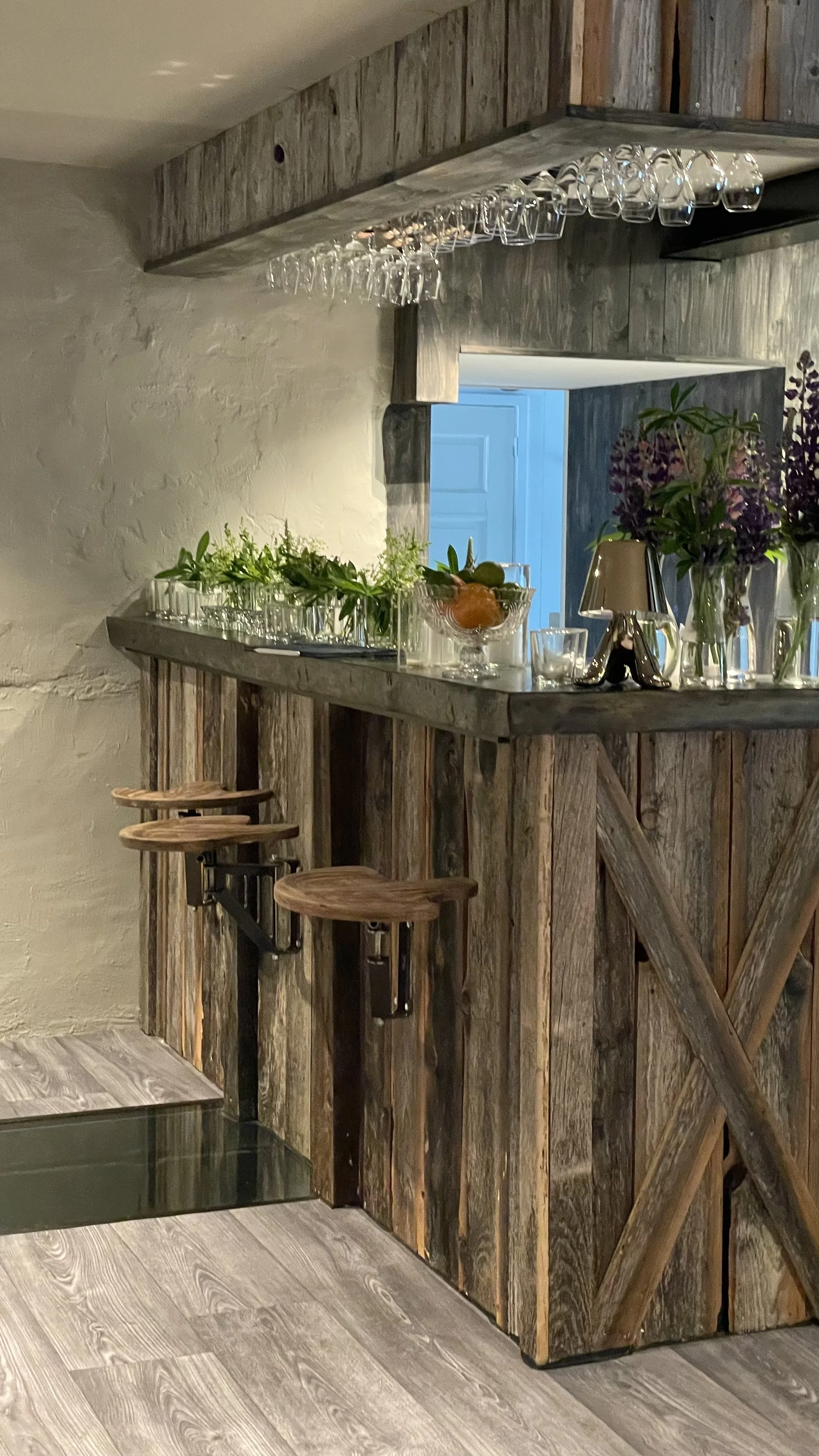 A rustic bar with a wooden countertop, three wooden bar stools, and various glassware and decorative items on the counter. A string of hanging wine glasses is above the bar, and there are plants and vases with flowers on the surface, with a view of a blue door in the background.