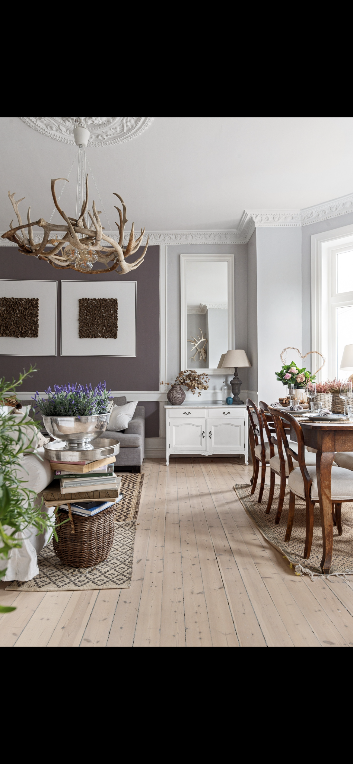 A cozy dining and living room area with a rustic chandelier made of antlers, a white sideboard with decor, a wooden dining table with chairs, and floral arrangements, featuring natural light from large windows and wood flooring.