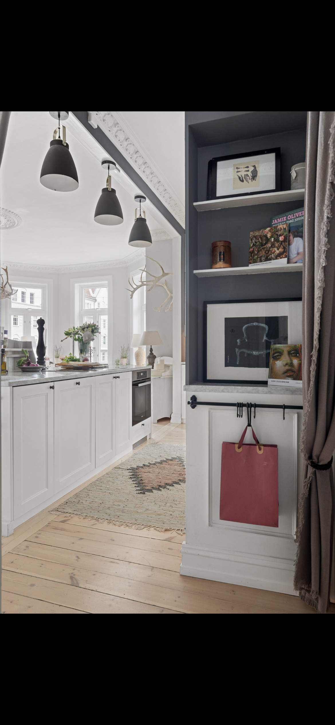Interior view of a bright kitchen and living space with white cabinets, a kitchen island, and decorative antlers on the wall. A built-in shelf displays framed pictures, books, and art. A pink shopping bag hangs on a black hook on a white paneled wall.