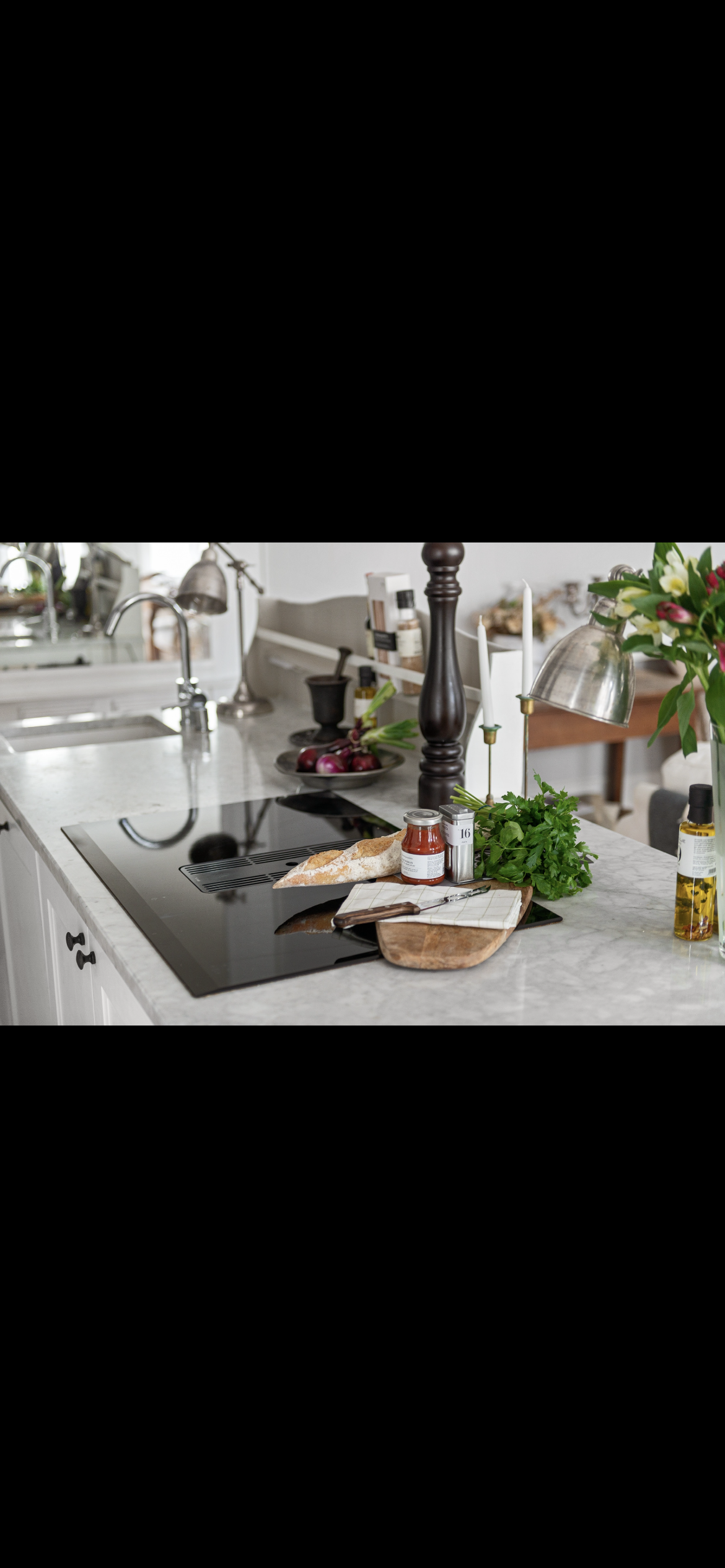 Kitchen countertop with a loaf of bread, a jar of tomato sauce, a cutting board, and fresh herbs, surrounded by various kitchen utensils and plants.