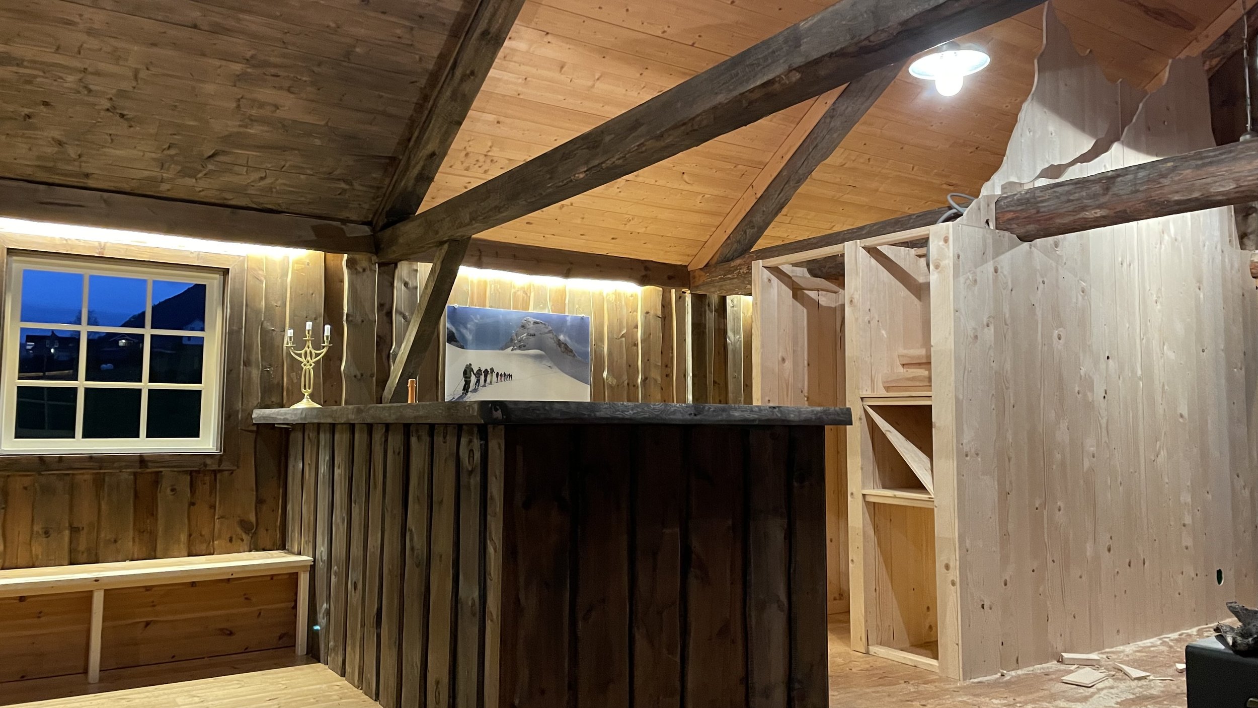 Interior of a wood-paneled room with a window, a picture of mountaineers on snow, a candelabra, and unfinished shelving.