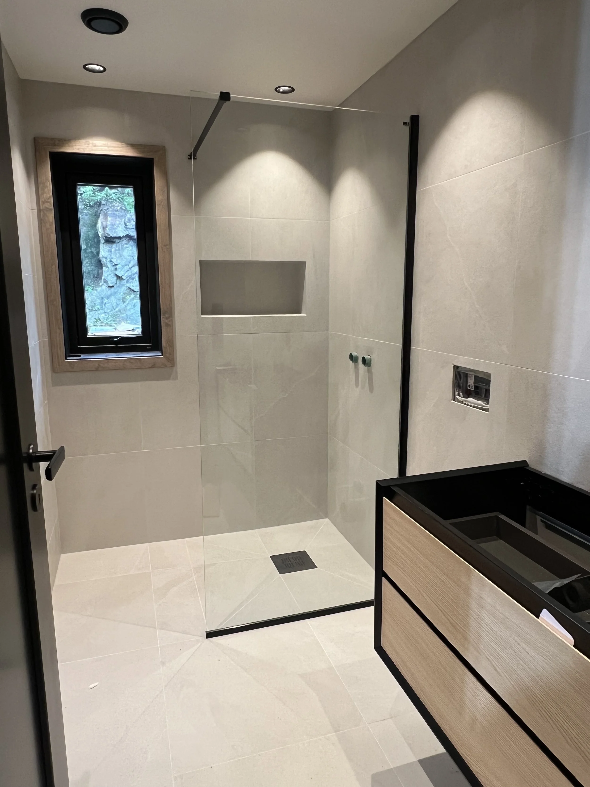 A modern bathroom with a walk-in shower featuring beige tiles, a small window with a wooden frame, built-in shelf in the shower, and a black vanity with drawers on the right side.