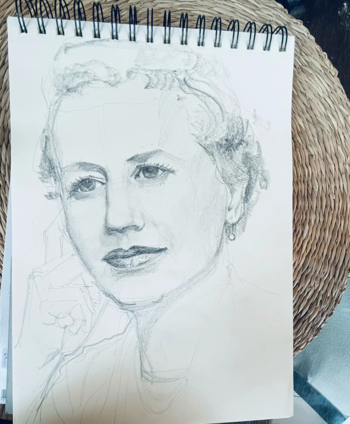A sketch of a woman's face in progress on a spiral-bound notebook, placed on a woven surface.