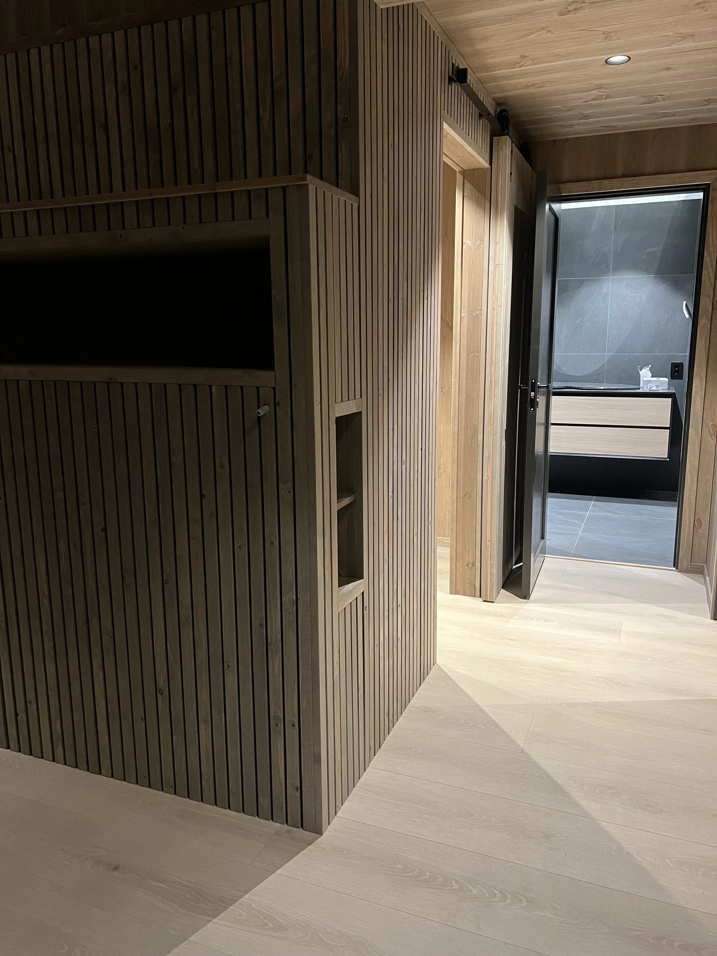Interior view of a modern building with wood-paneled walls and flooring, showing a doorway leading to a bathroom with dark tiles and a wooden vanity.
