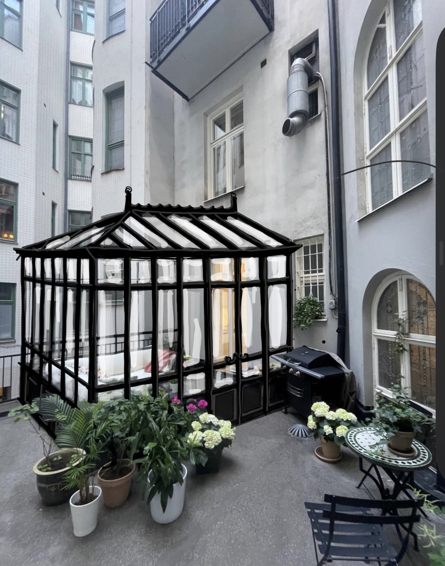 An outdoor city balcony with potted plants, including flowers and greenery, a small round table with a potted plant, a black grill, and a black-framed glass conservatory structure with a peaked roof, set against tall apartment buildings with windows and balconies.