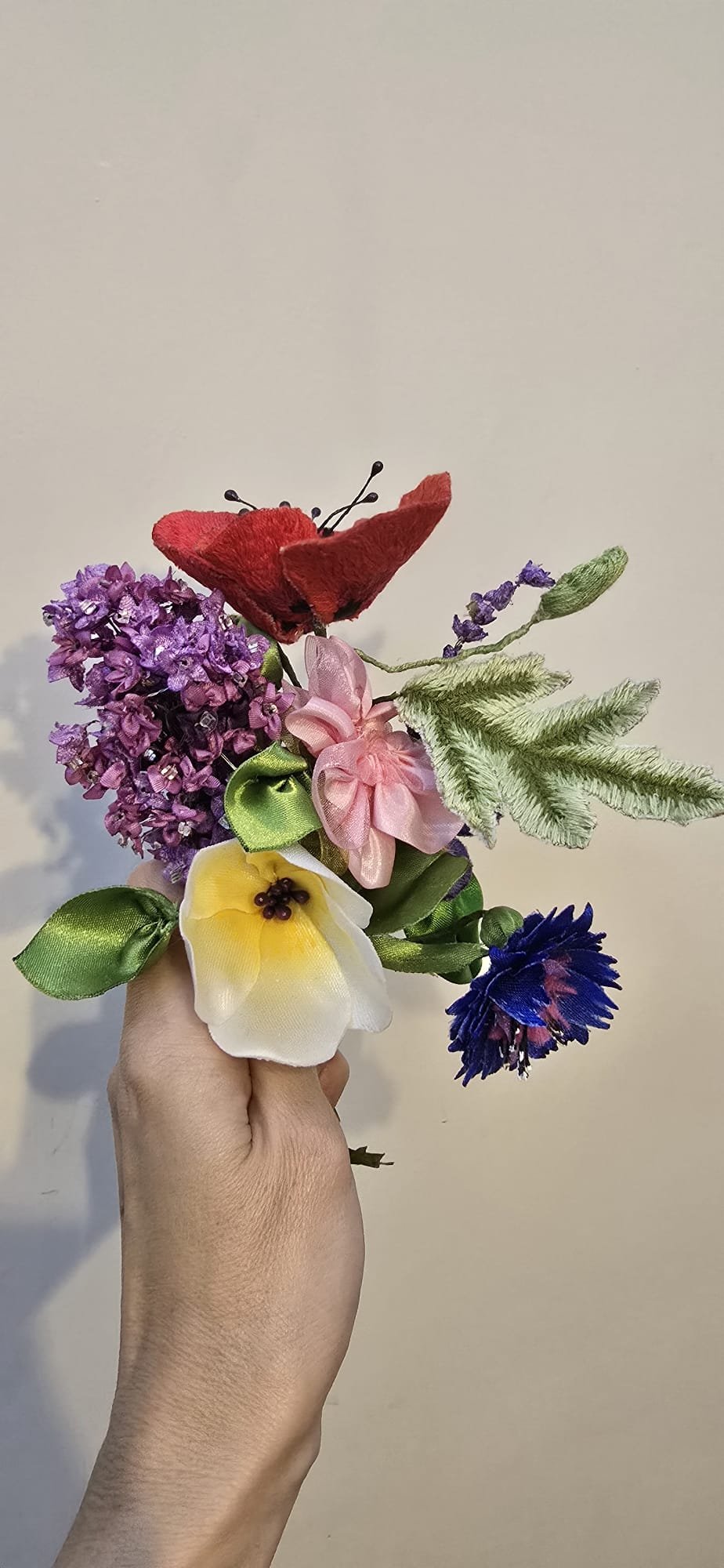 A hand holding a small bouquet of artificial flowers, including a red butterfly, purple lilac, pink ribbon flower, yellow daffodil, and blue cornflower.