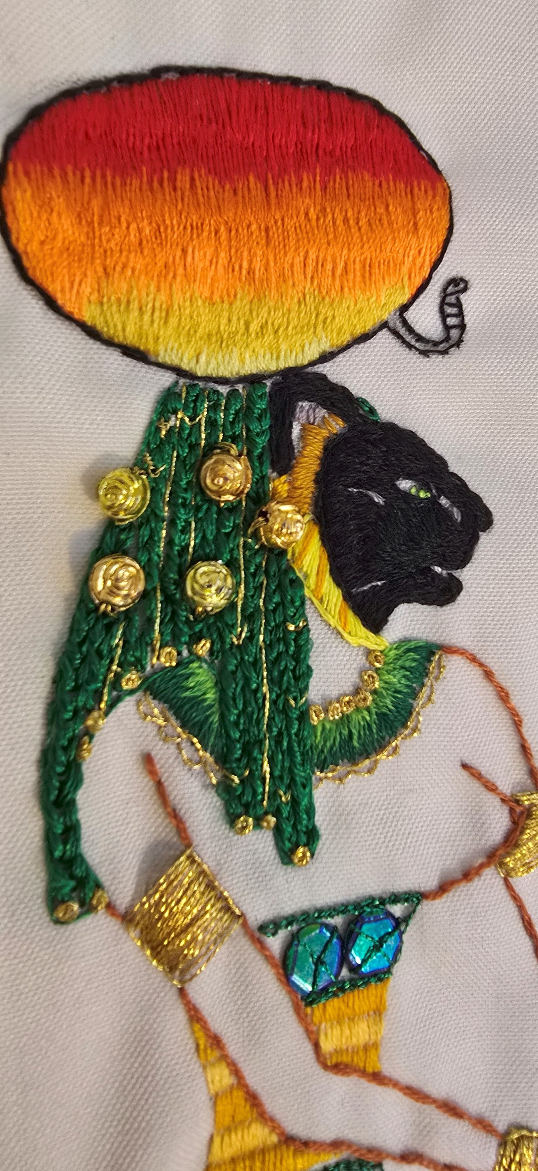 Embroidery artwork of a person with a rainbow-colored head, black face, green attire with gold embellishments, and a yellow garment.