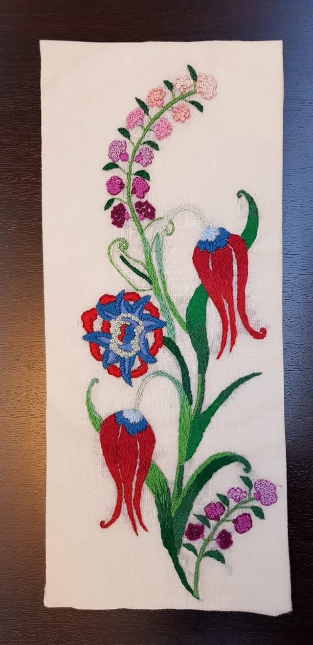 Embroidery of a flower with purple, pink, and red blossoms, blue and red accents, and green leaves on white fabric.