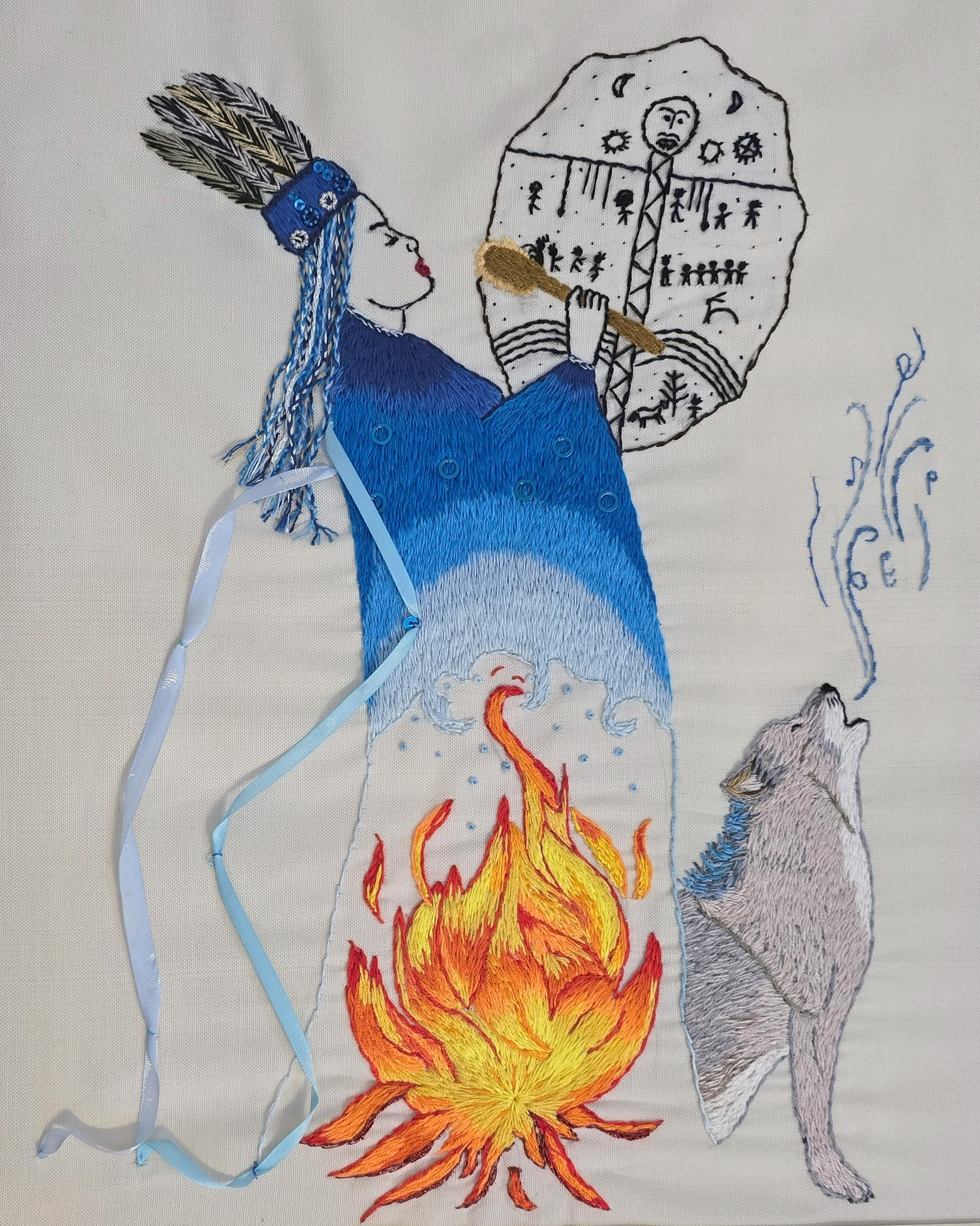 Embroidery artwork depicting a woman with long hair and a headdress, holding a makeshift drum or shield, with a wolf howling nearby and flames below. The design combines stitched and fabric elements with blue and gray tones.