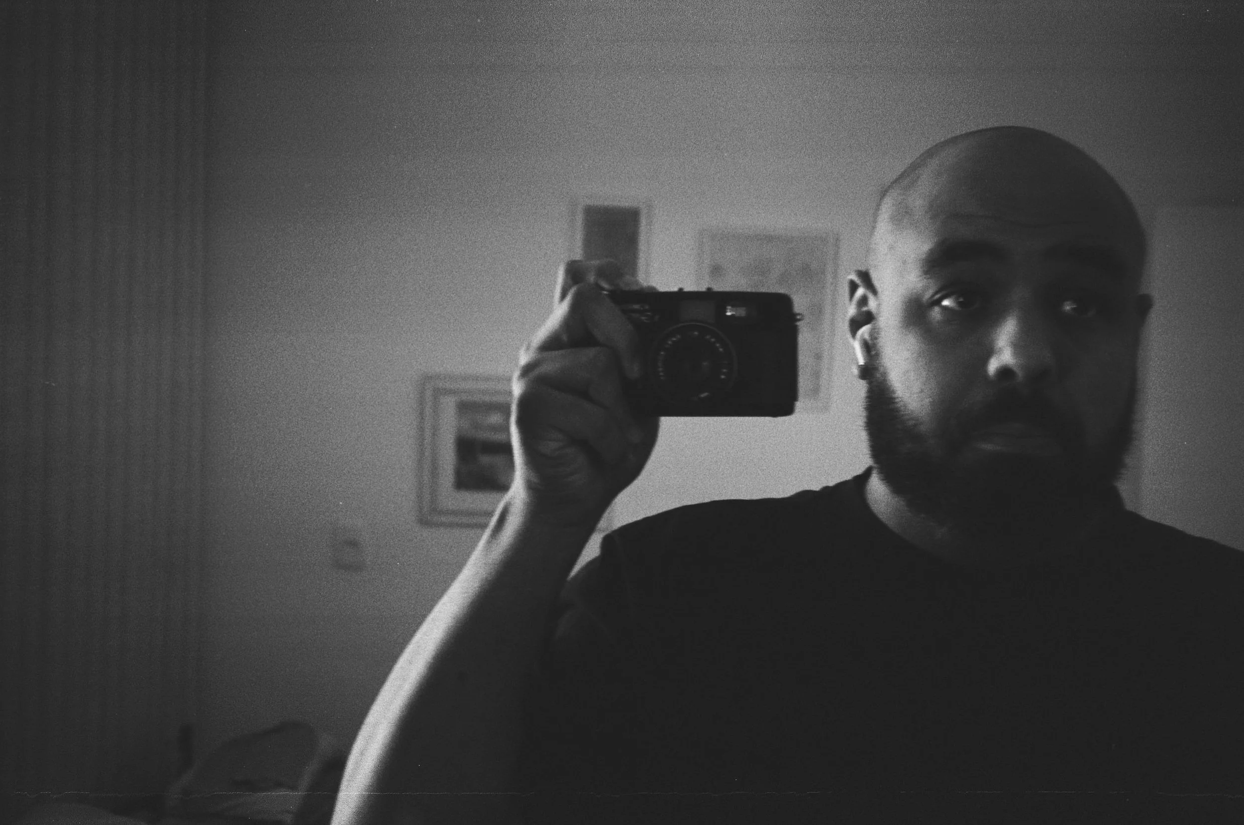 Homem com barba e cabelo raspado, segurando uma câmera fotográfica na frente de espelho, em um ambiente com fotos na parede ao fundo, em preto e branco.
