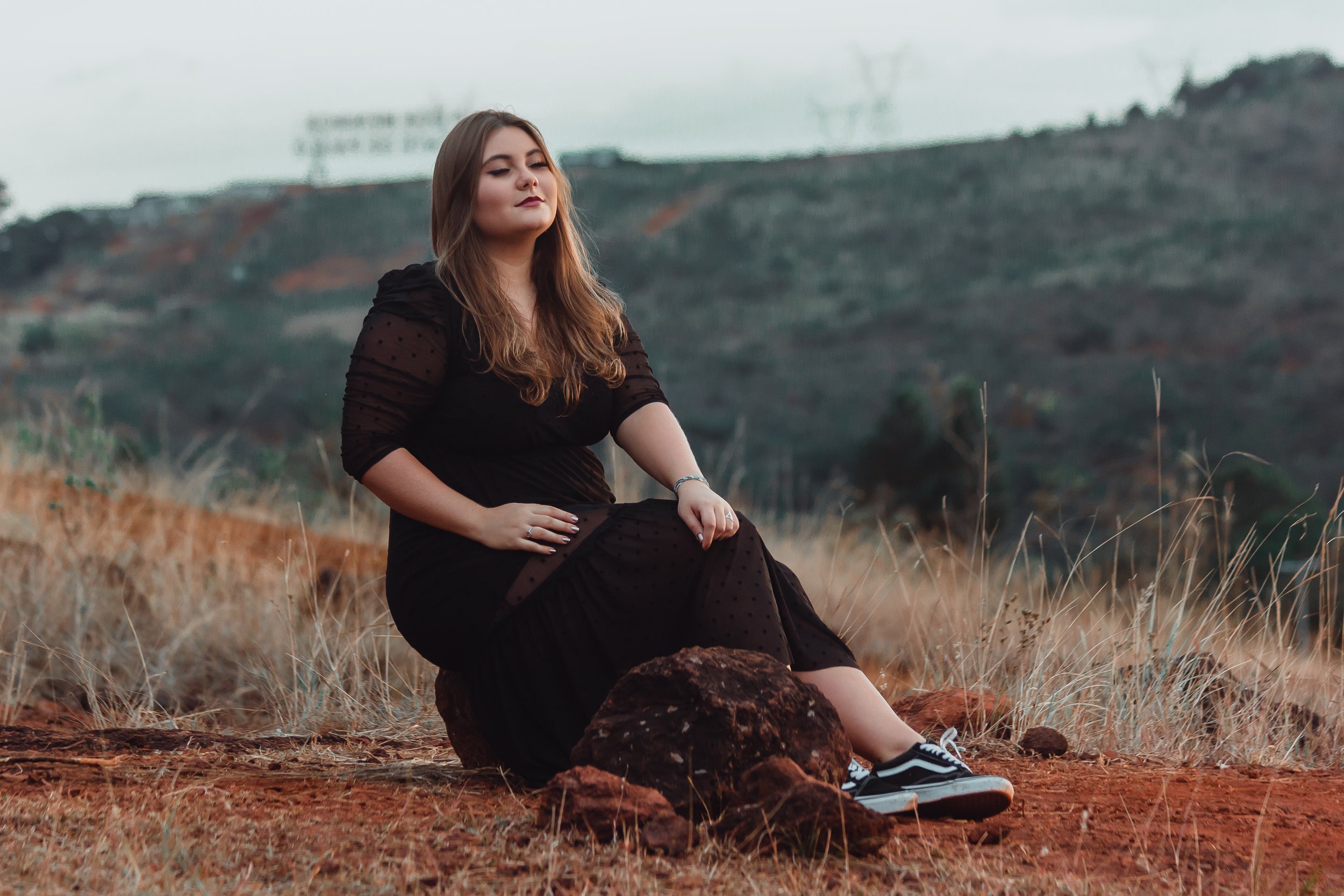 Mari Vieira sentada sobre uma pedra de olhos fechados, com vestido preto longo e tênis, diante da paisagem de serra no Topo do Mundo, ensaio fotográfico em Belo Horizonte