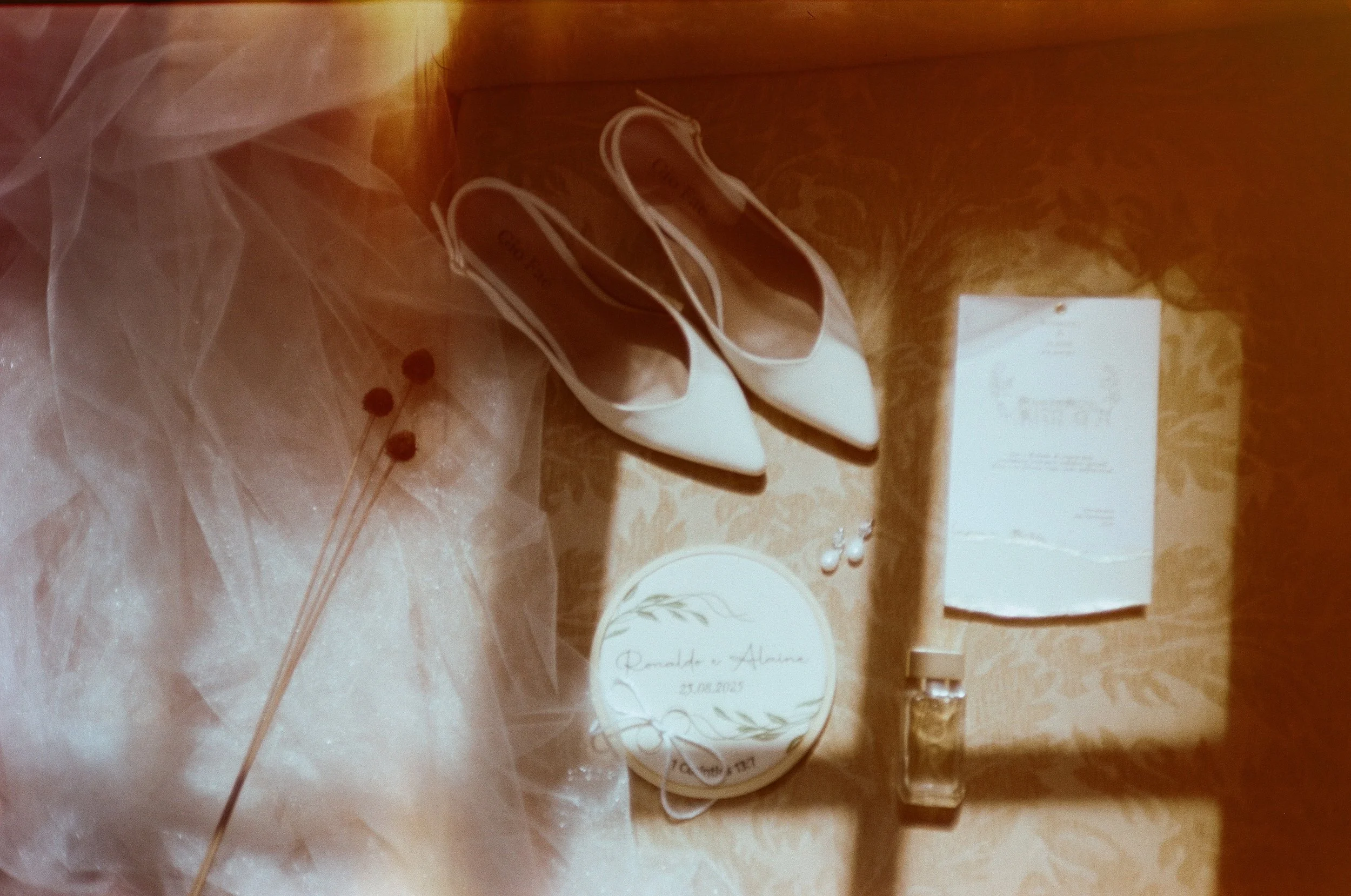 Detalhes da noiva sobre a cama: sapatos brancos, véu, convite, caixa personalizada com os nomes dos noivos, brincos e perfume, no casamento de Alaine e Ronaldo em Belo Horizonte, fotografia analógica