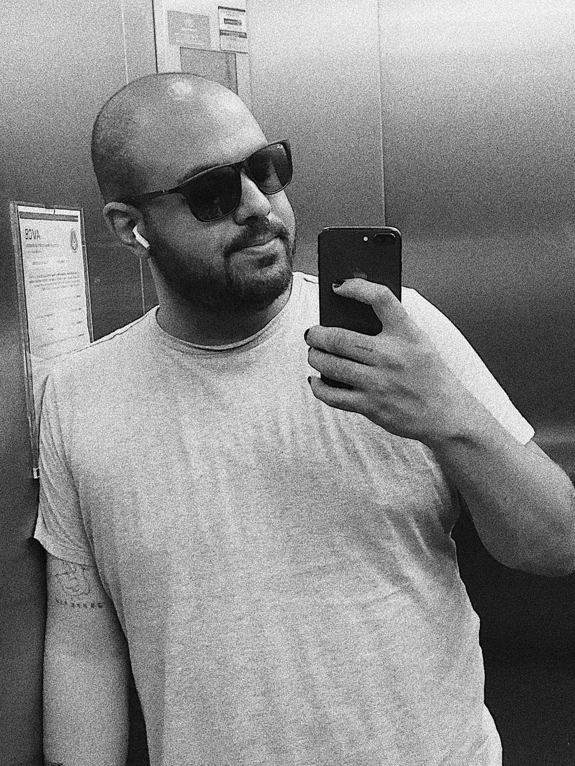 Homem de cabeça raspada, usando óculos escuros, camiseta cinza, ouvindo fones de ouvido, tirando selfie com smartphone em um elevador